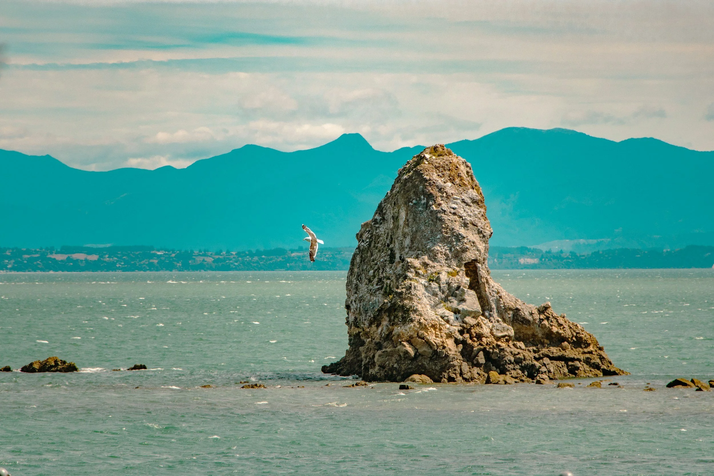 A large rock formation in the ocean with smaller rocks nearby. In the background, there are mountain ranges under a partly cloudy sky. A bird is flying near the large rock in the foreground.