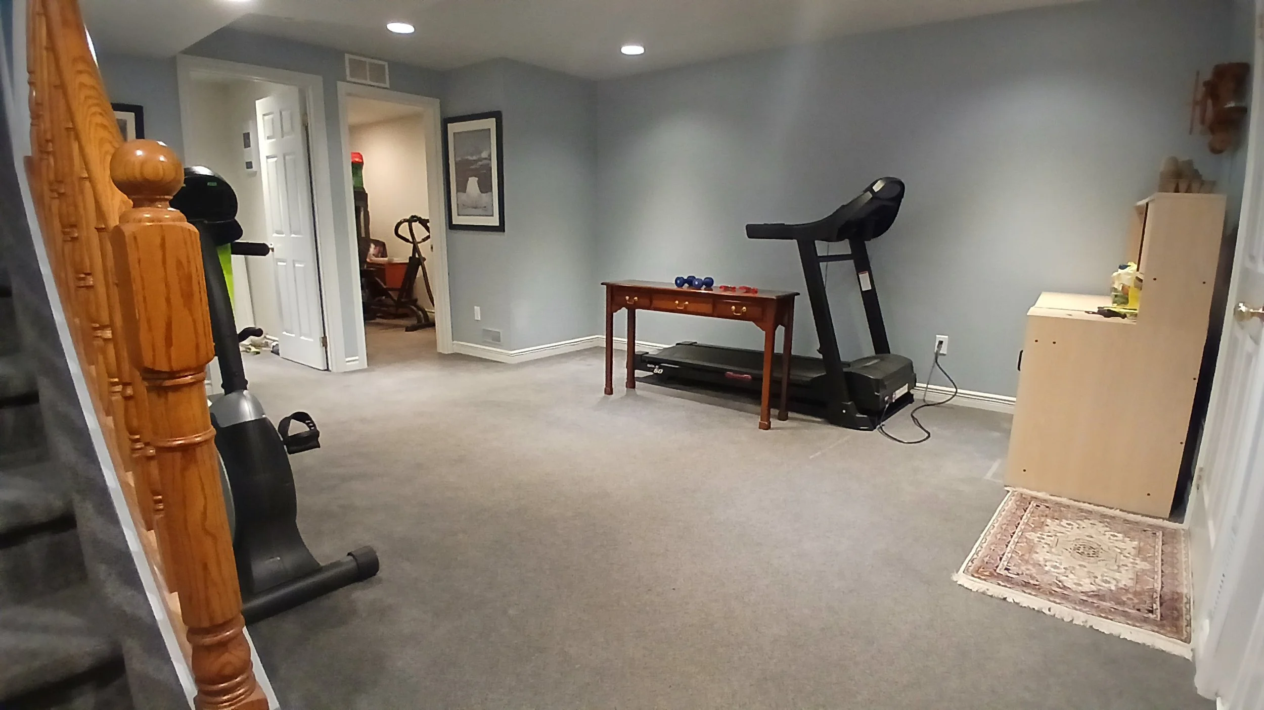 A living room corner with exercise equipment, including a treadmill and a desk with small dumbbells, beige carpet, and a small rug near a staircase with wooden banisters.