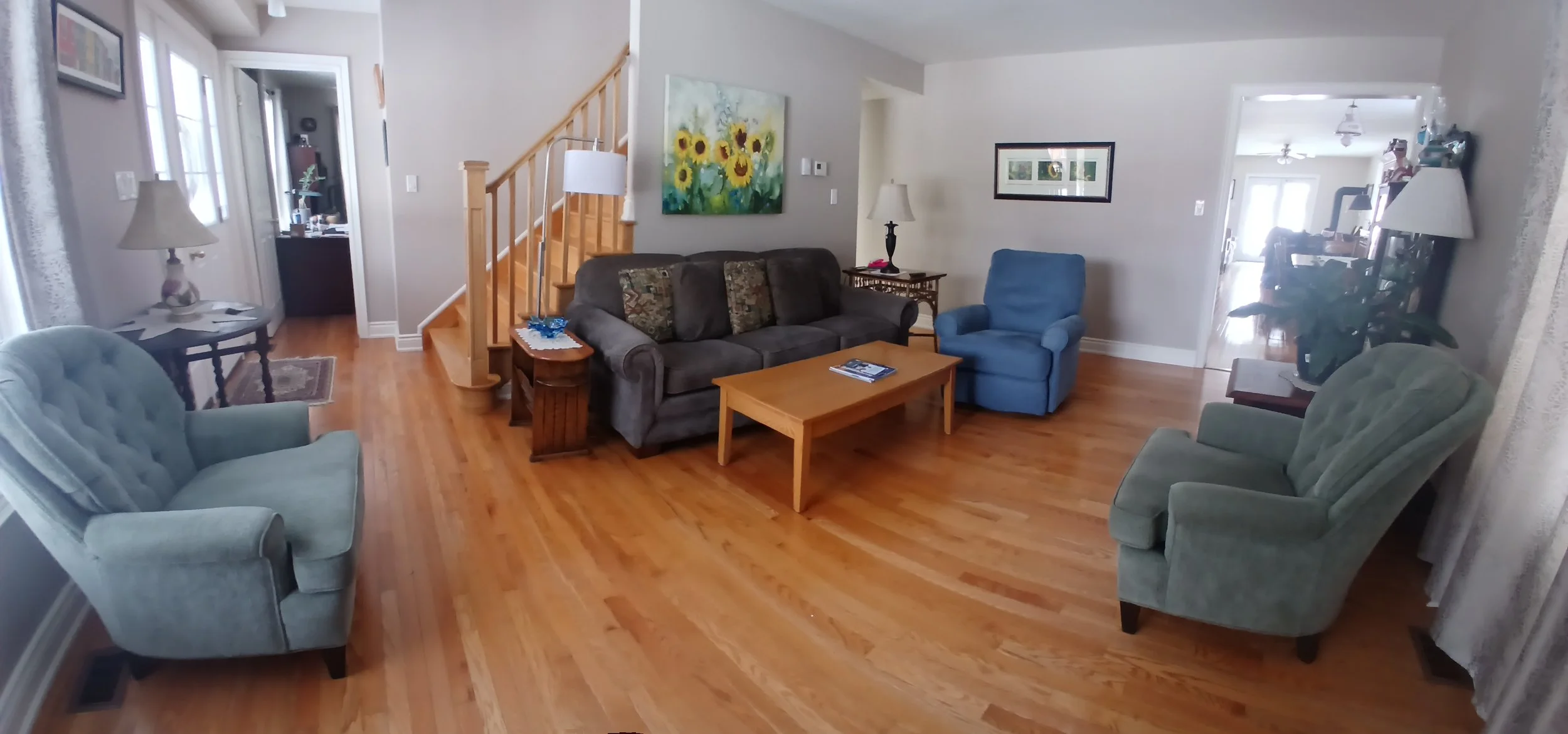 Living room with hardwood floors, a gray sofa, blue armchair, green armchairs, wooden coffee table, side tables with lamps, wall art, and staircase.