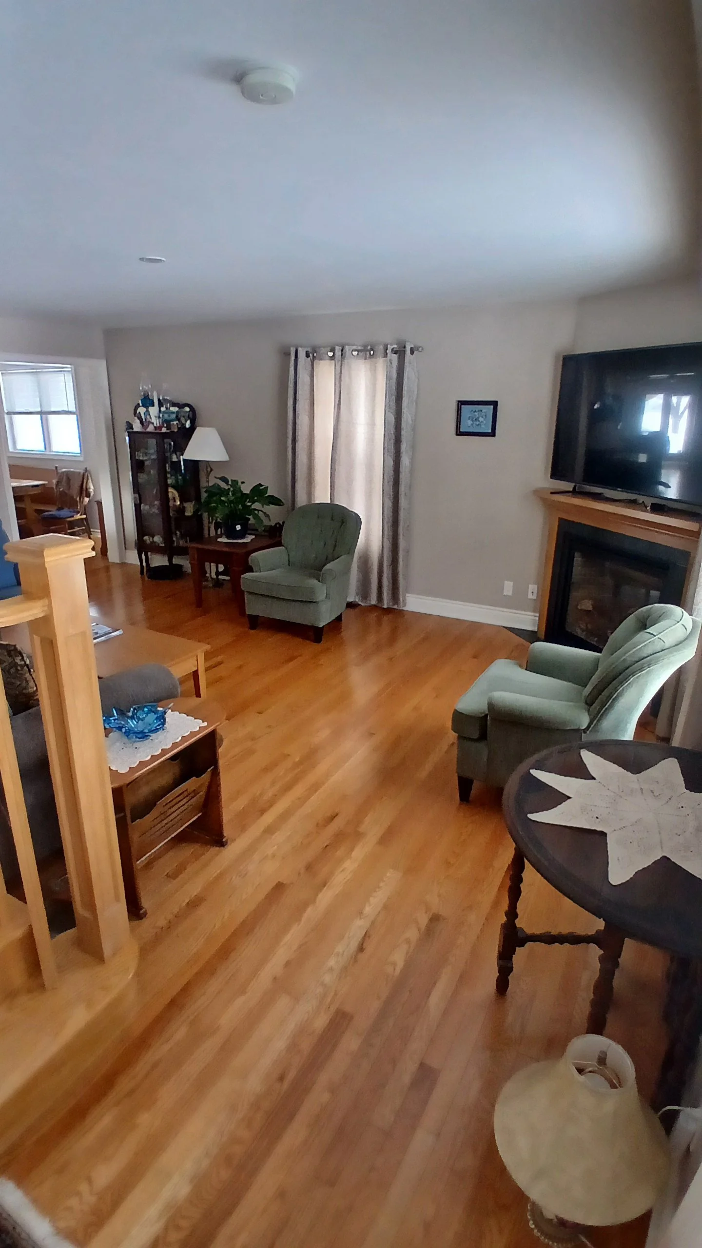 Living room with hardwood floor, green armchairs, a television over a fireplace, a curtain covering a window, and a small table with a leaf-shaped tablecloth.