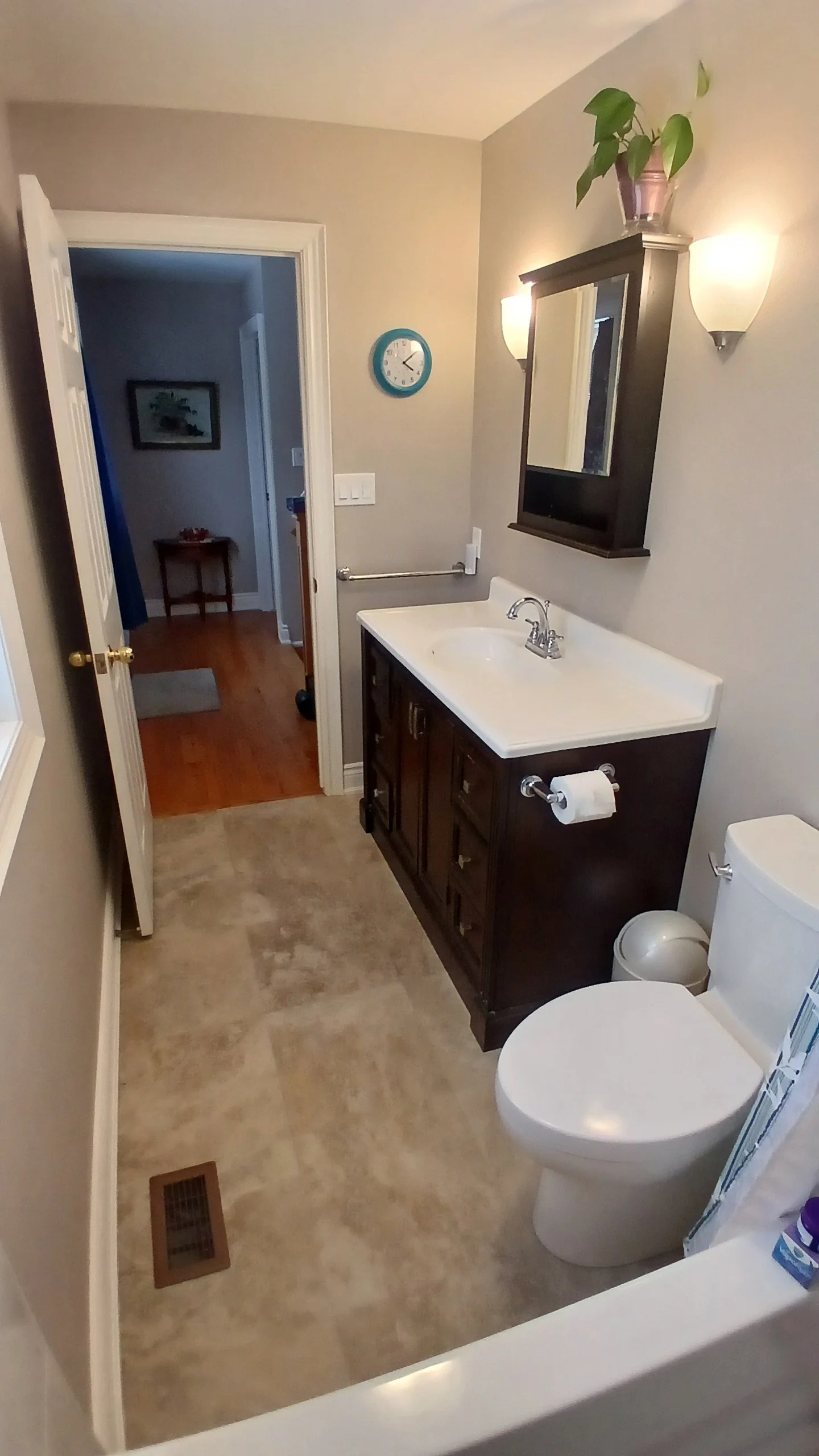 A small bathroom with a dark wood vanity, white sink, beige walls, a wall mirror, wall sconces, a potted plant on top of the mirror cabinet, and a toilet with toilet paper. The floor is beige tile, and there is a vent near the floor. An open door lea