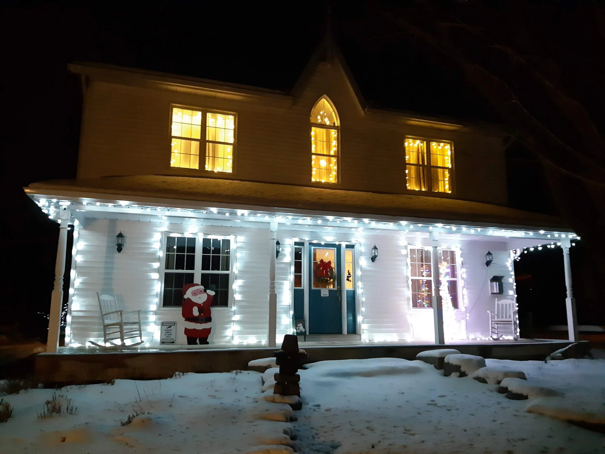 A house decorated for Christmas with yellow lights in the windows, a Santa Claus figure outside, and snow on the ground.