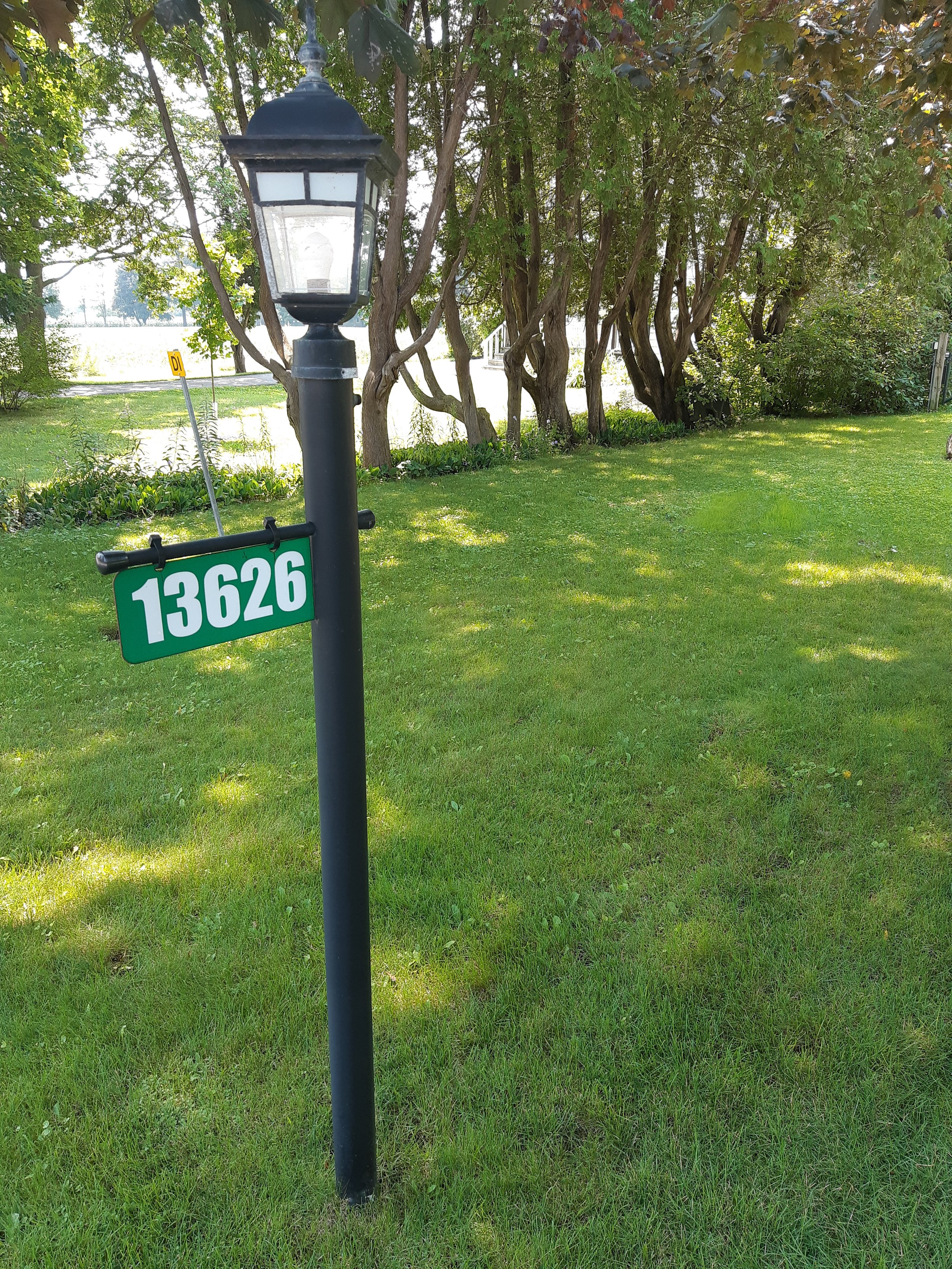 A black lamp post with a green address sign that has the number 13626 on it, in a grassy yard with trees and bushes in the background.