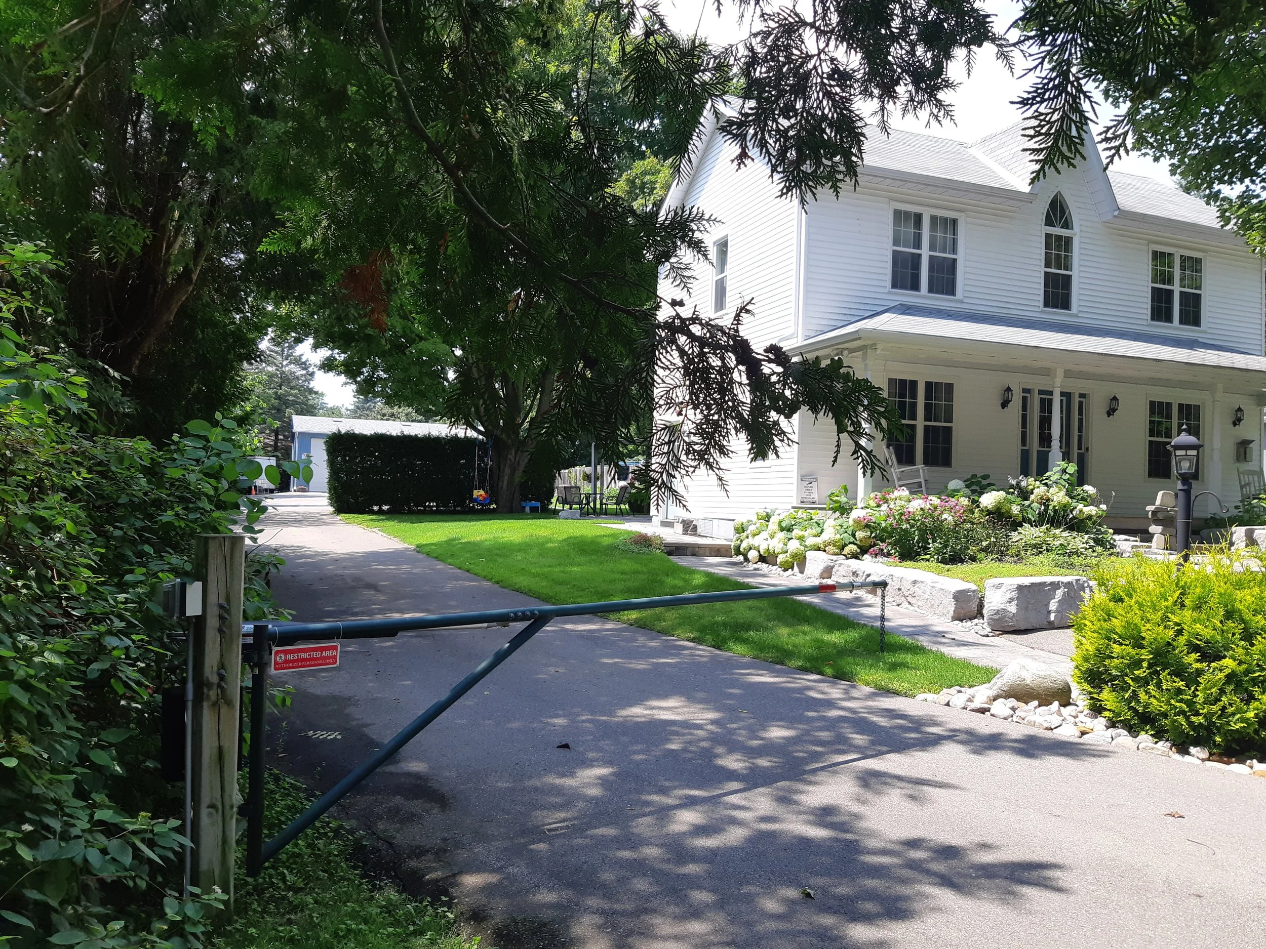 A white two-story house with multiple windows, surrounded by well-maintained greenery and colorful flowers, partially obscured by trees, with a driveway and a small gate at the front.