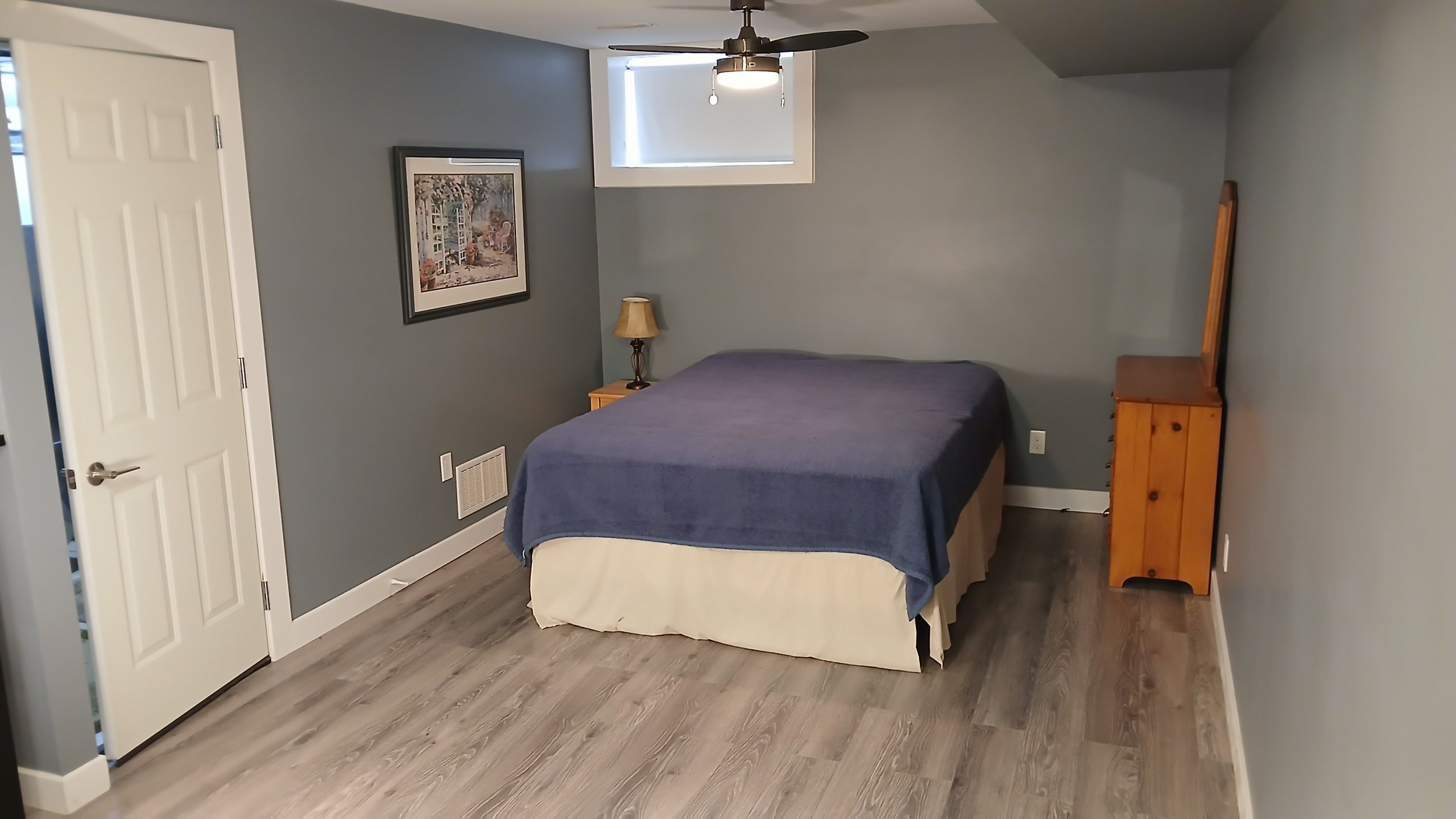 A small bedroom with a bed covered in a blue blanket and a white skirt, a wooden nightstand with a lamp, a framed picture on the wall, a window with blinds, a ceiling fan with a light, and a wooden dresser.
