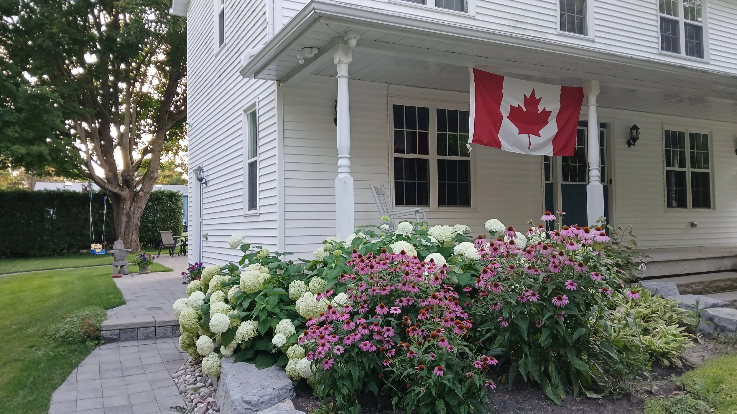 White house with a porch hanging a Canadian flag, surrounded by flowers and greenery.