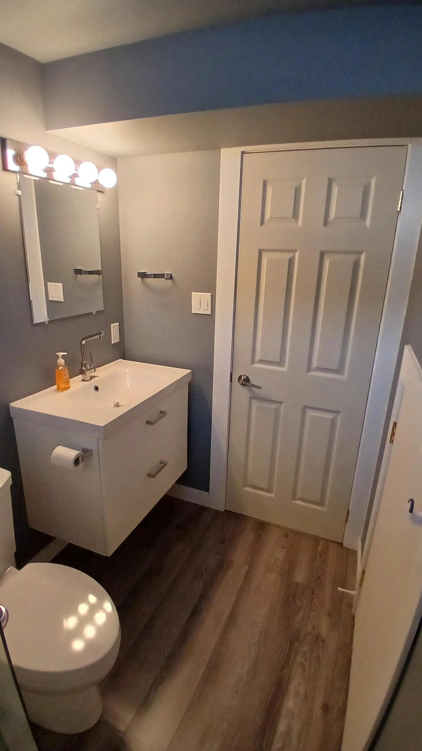 A small bathroom with a white sink and cabinet, a mirror with lights above, a toilet, and a closed door. The walls are painted gray, and the floor is wood-like.