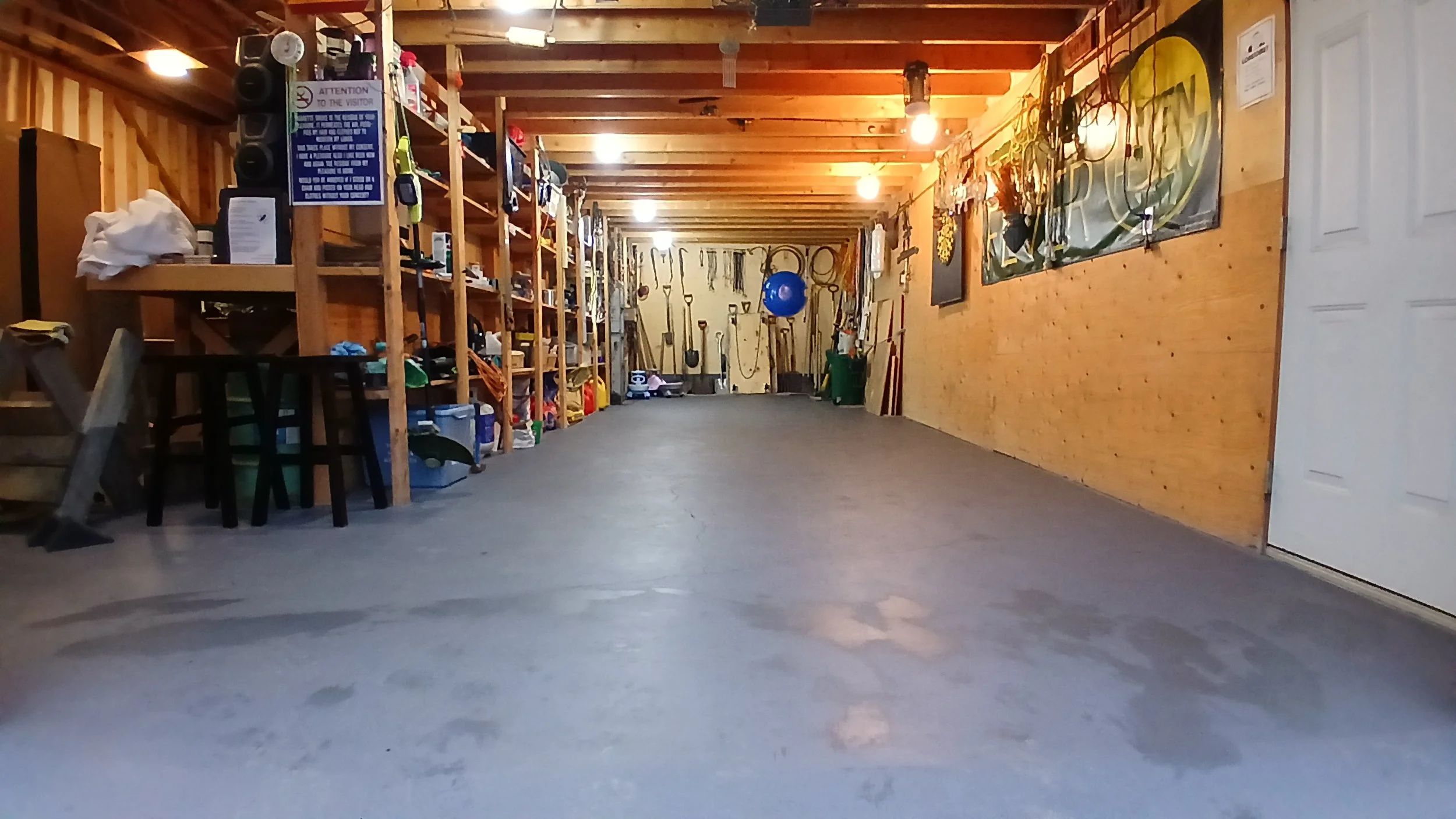 A garage with wooden walls and ceiling, concrete floor, guitar and sports equipment on shelves, a blue exercise ball hanging, and garage tools on the back wall.