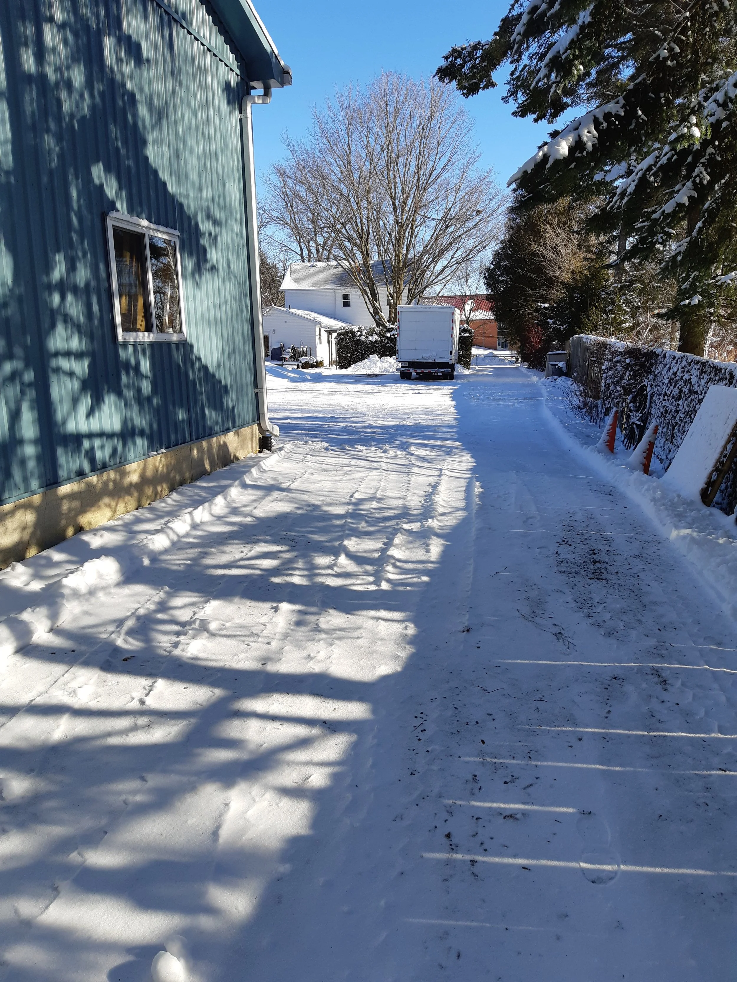 Snow-covered residential driveway with a blue house on the left, a large leafless tree in the background, a white truck parked in the distance, and snow-covered trees on the right side. Shadows cast on the snow.