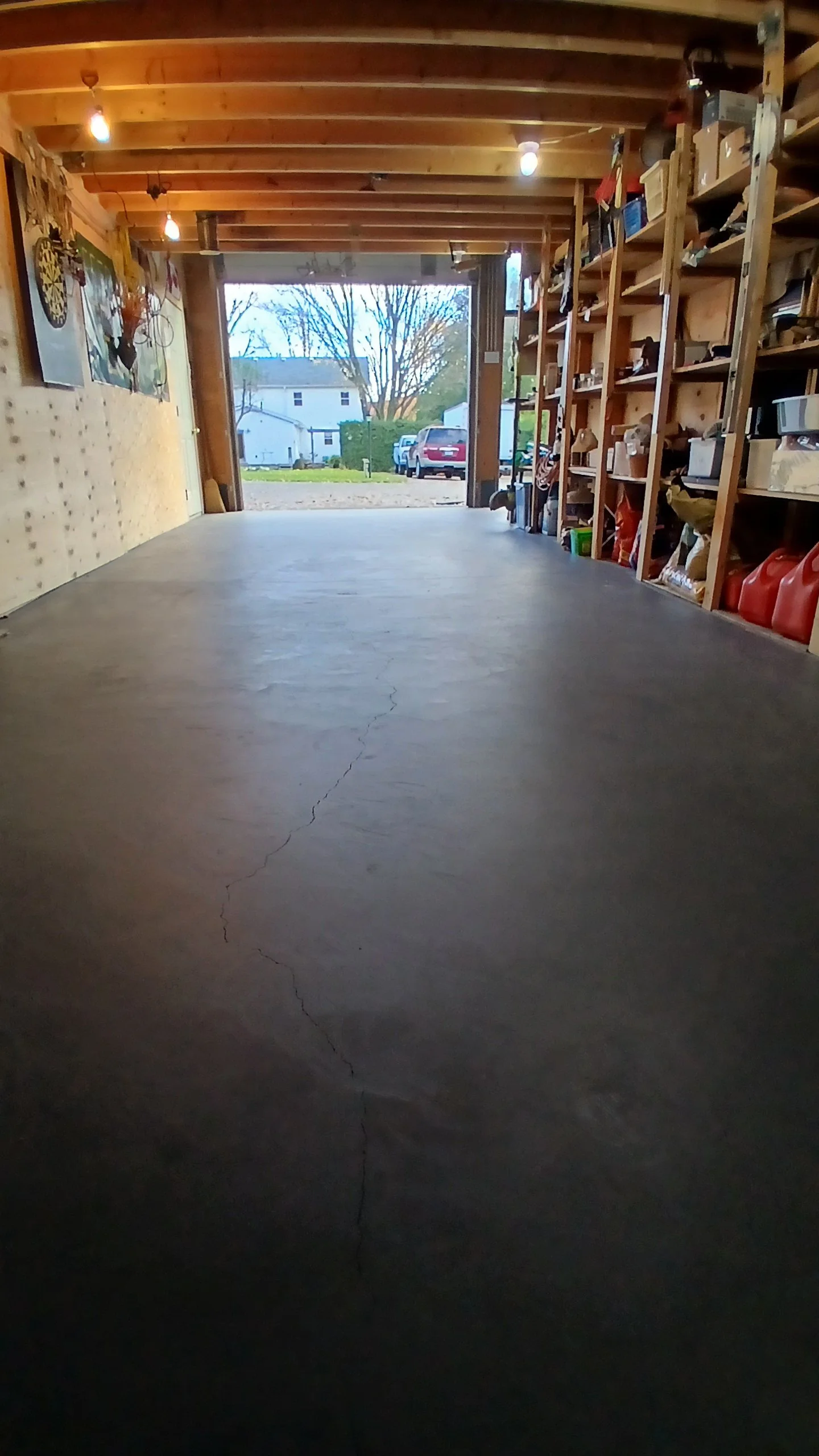 View of a garage interior with a concrete floor, wooden ceiling and shelves on the right filled with various items, open garage door showing a driveway yard, parked cars, and neighboring houses outside.