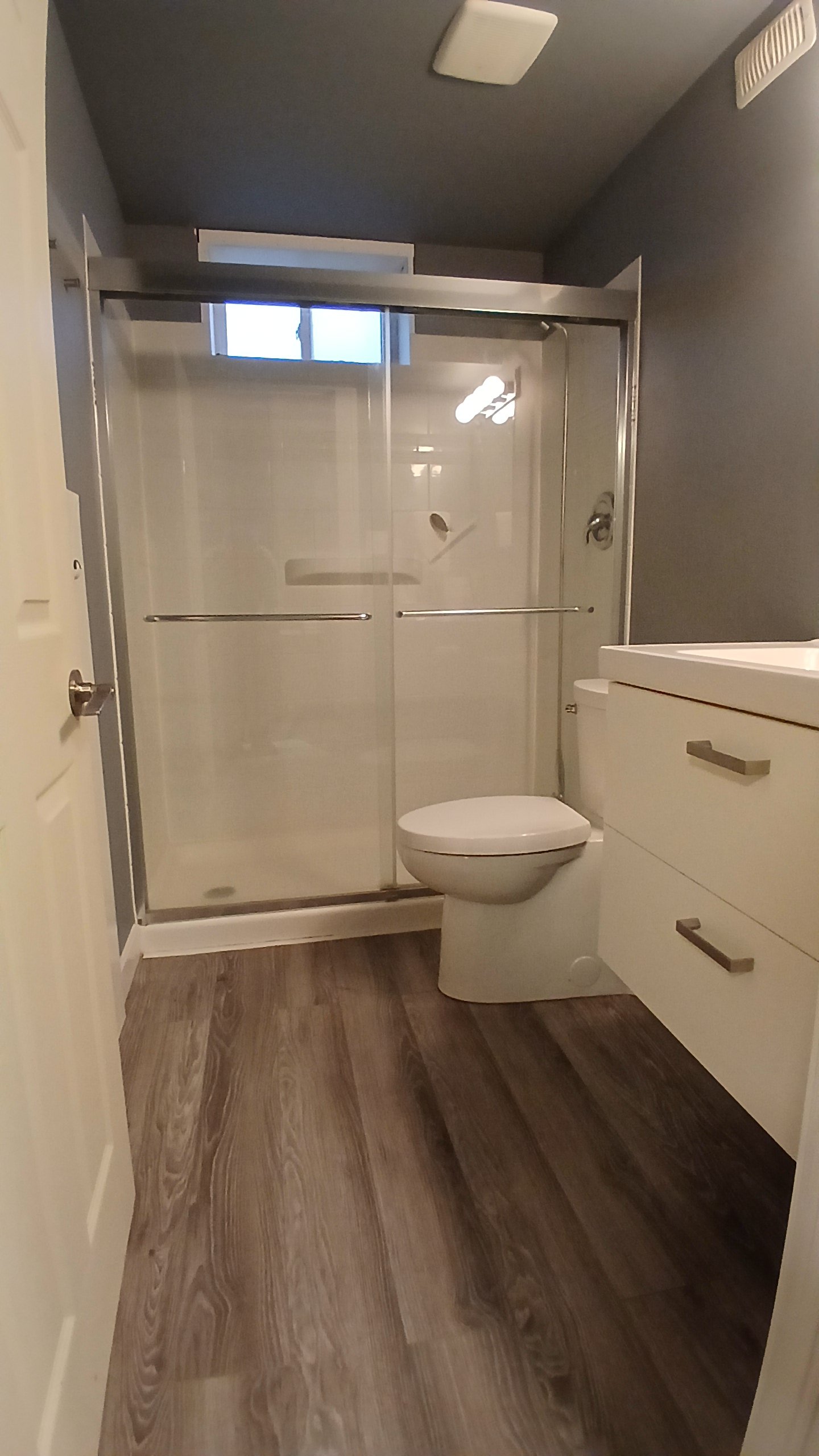 A bathroom with a shower stall with glass sliding doors, a toilet, and a white vanity with drawers. The floor is wood-style flooring, and the walls are painted gray. There is a small window above the shower.