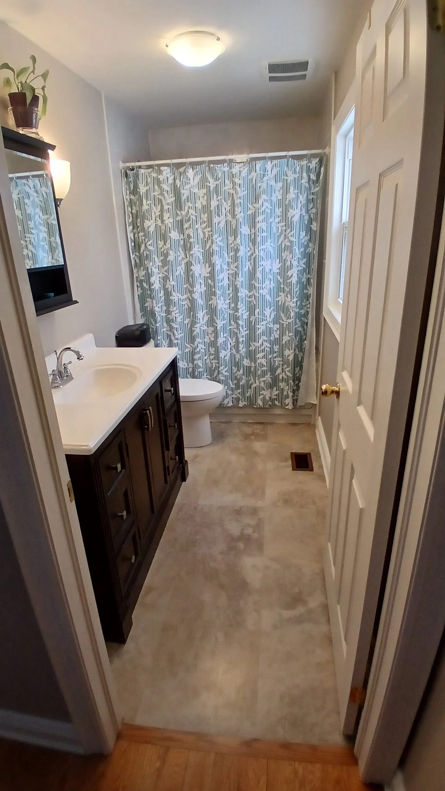 A small bathroom with light walls, a dark wooden vanity with a white sink, a mirror, a small potted plant, a sink soap dispenser, a toilet, a shower with patterned curtains, a window, an overhead light, and a vent in the ceiling.
