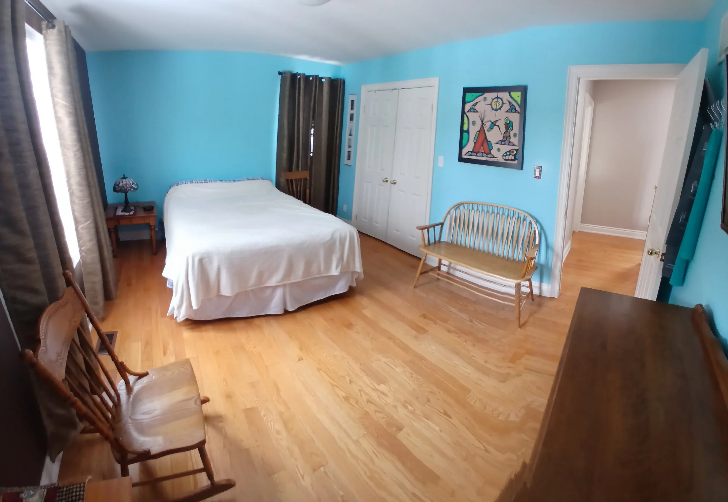 A bedroom with blue walls, hardwood floors, a bed with a white cover, a nightstand with a lamp and a phone, a wooden bench, and decor including a colorful abstract painting and framed photos.