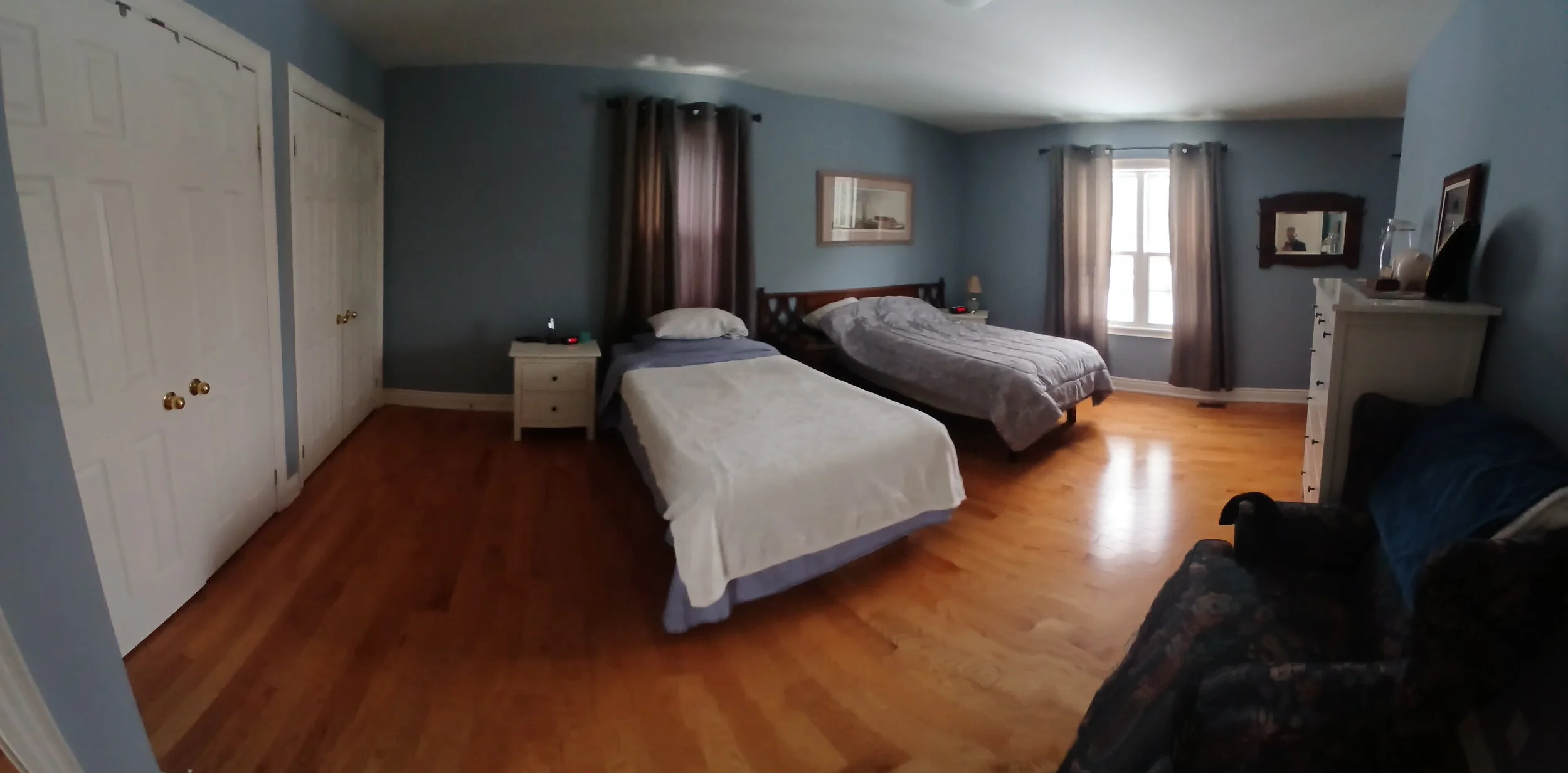 A bedroom with two beds, a dresser, and a window with curtains, featuring wooden flooring and light blue walls.