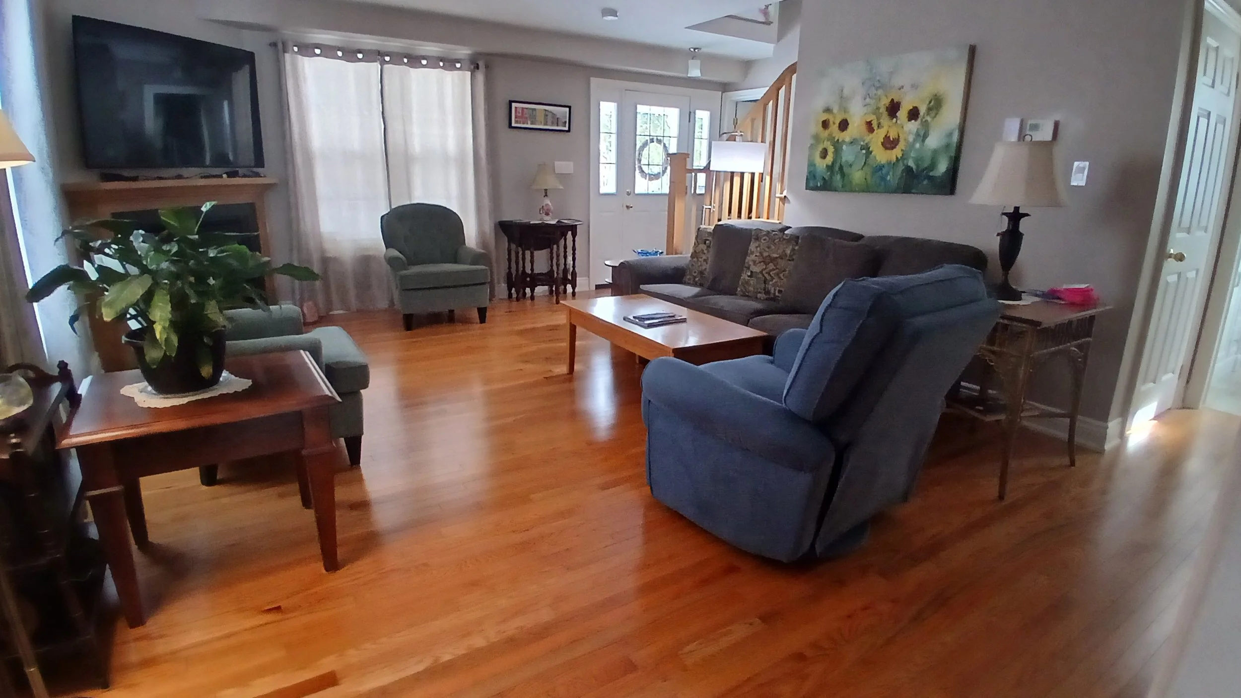Living room with a large TV mounted on the wall, a green armchair, a wooden coffee table, blue and patterned upholstered chairs, a floor lamp, a side table with a lamp, a painting of sunflowers, windows with curtains, and hardwood floors.