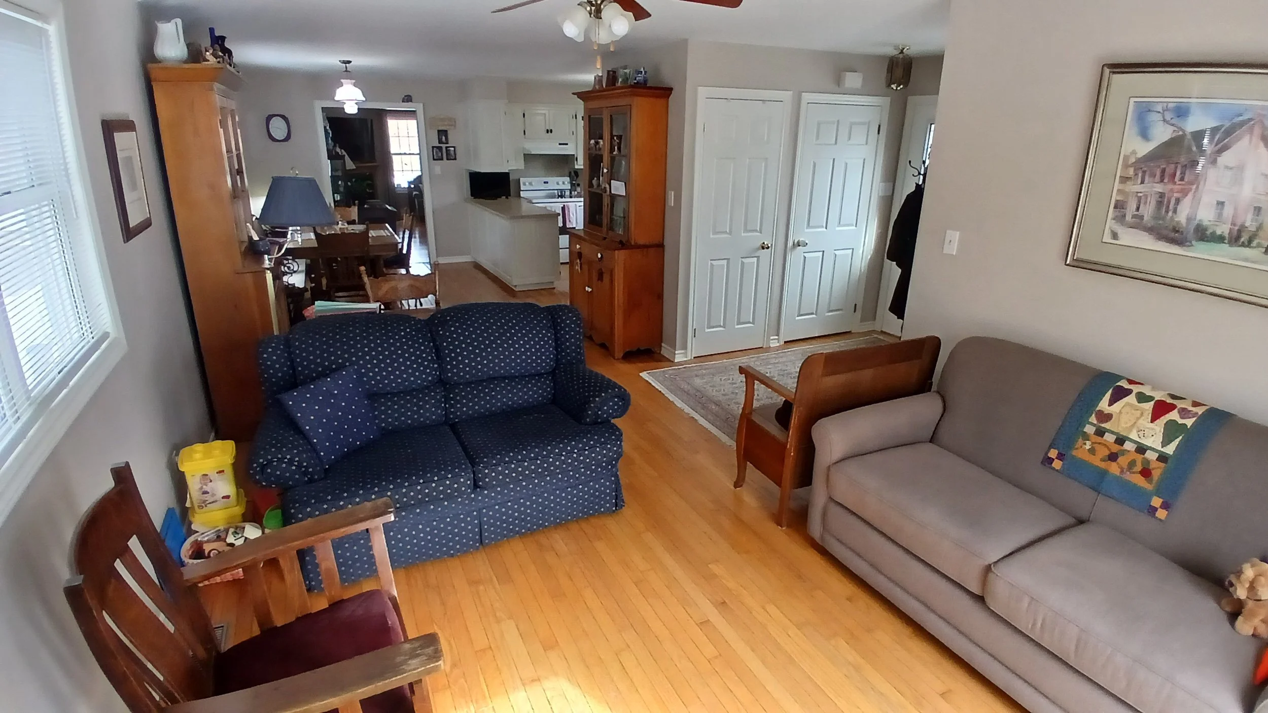 Living room with a beige sofa and a navy blue sofa with polka dots, a wooden chair with a quilt draped over it, a small wooden table, and hardwood floors. In the background, a dining area with a dining table, chairs, a china cabinet, and kitchen visi