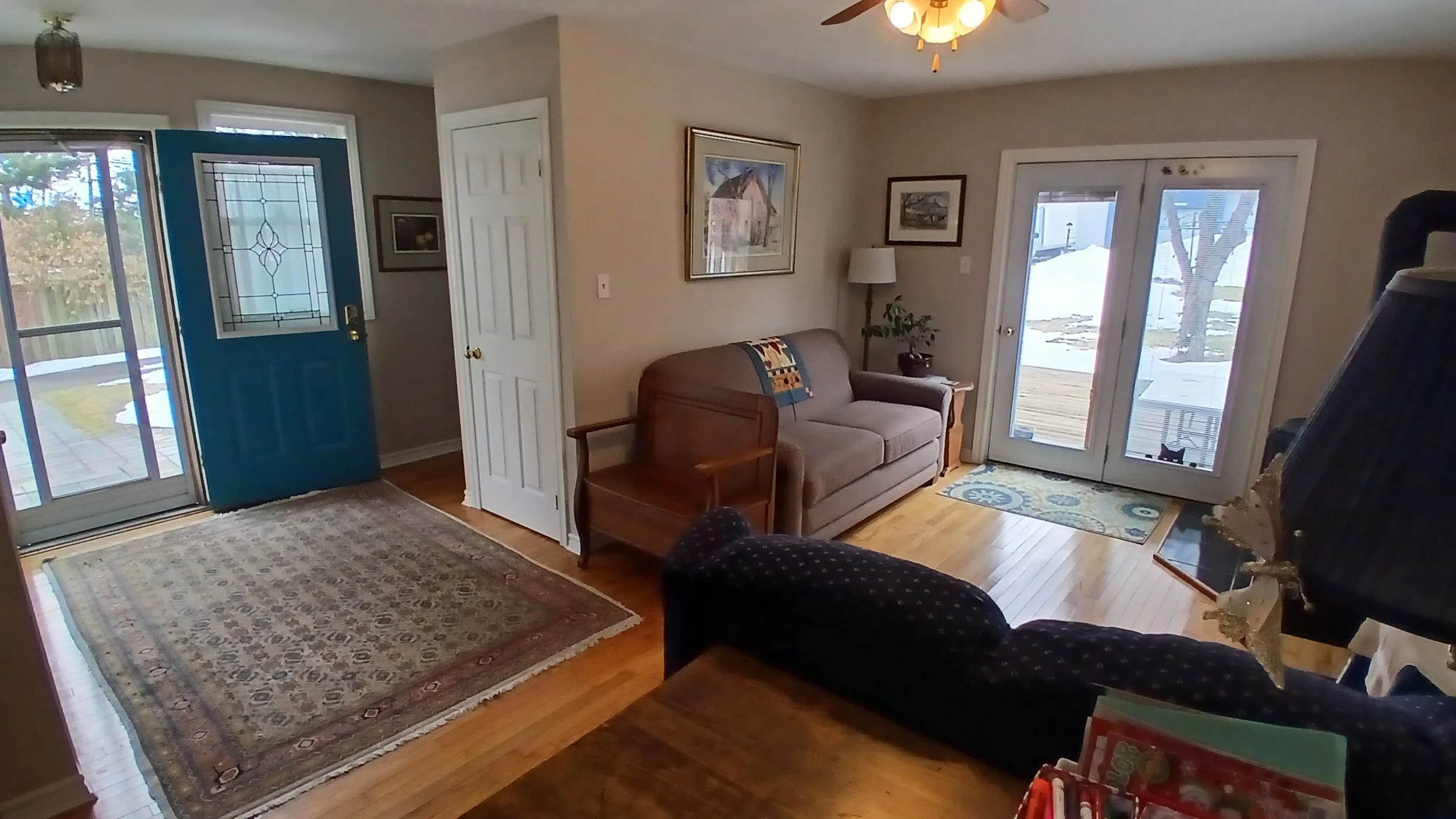 Living room with hardwood floors, a patterned rug, a beige couch with a colorful blanket, two paintings on the wall, a wooden chair, a floral mat by glass patio doors, and a ceiling fan with lights.