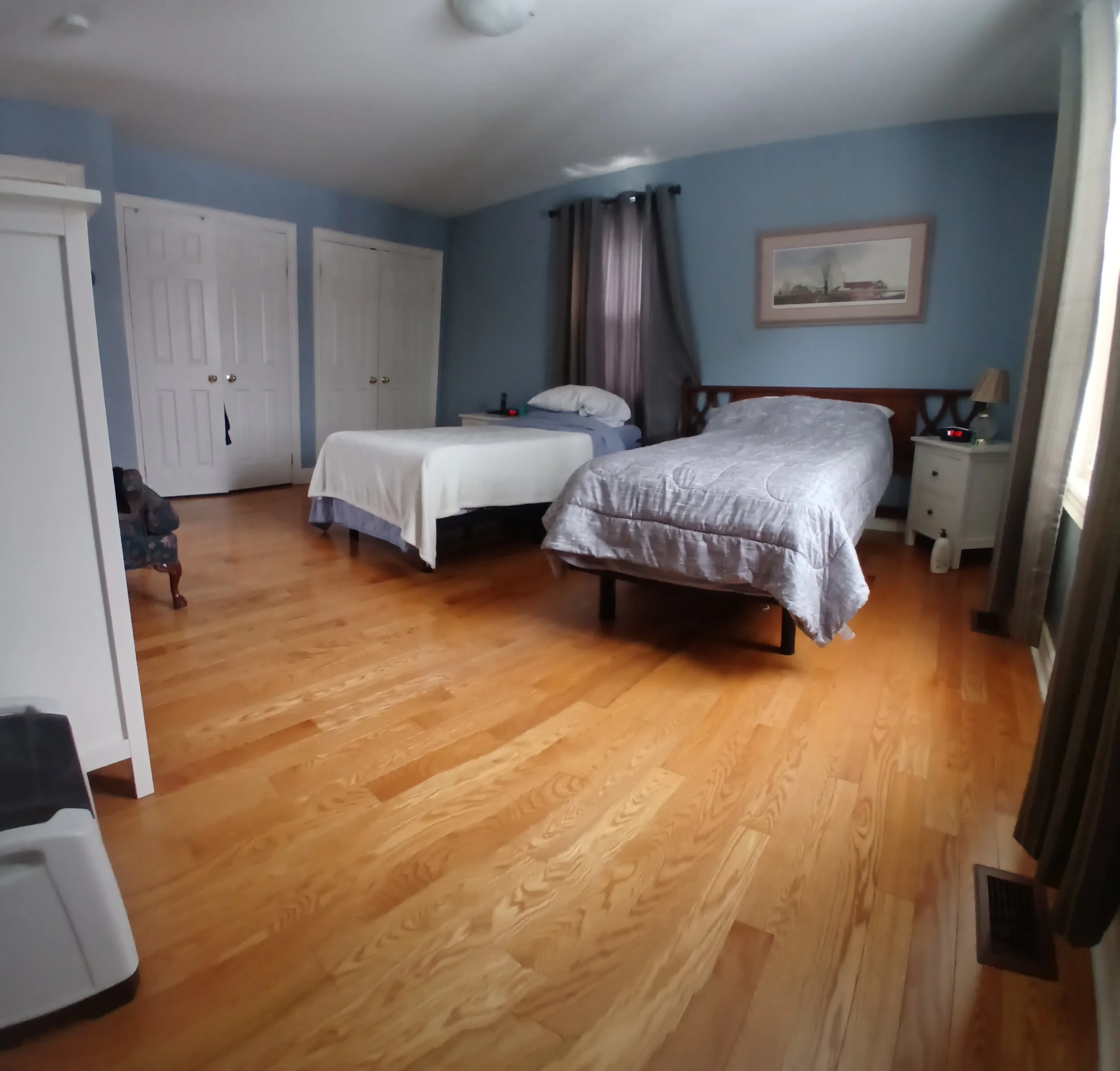 A bedroom with light blue walls, wooden flooring, two beds with beige and gray bedspreads, a nightstand with a lamp and clock, a dresser, a framed picture on the wall, and a window with dark curtains.