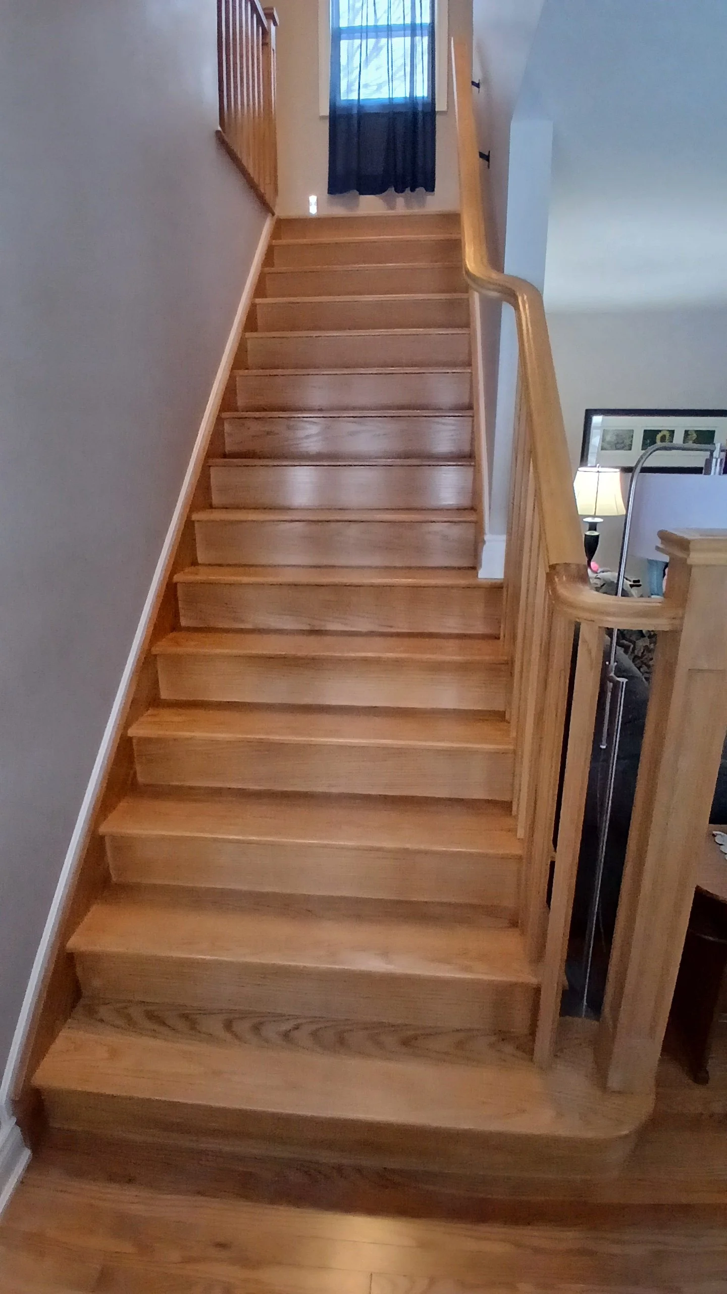 Wooden staircase with a curved handrail leading to an upper floor, a window with a sheer curtain at the top of the stairs, and part of a room with a lamp and framed pictures visible on the right.