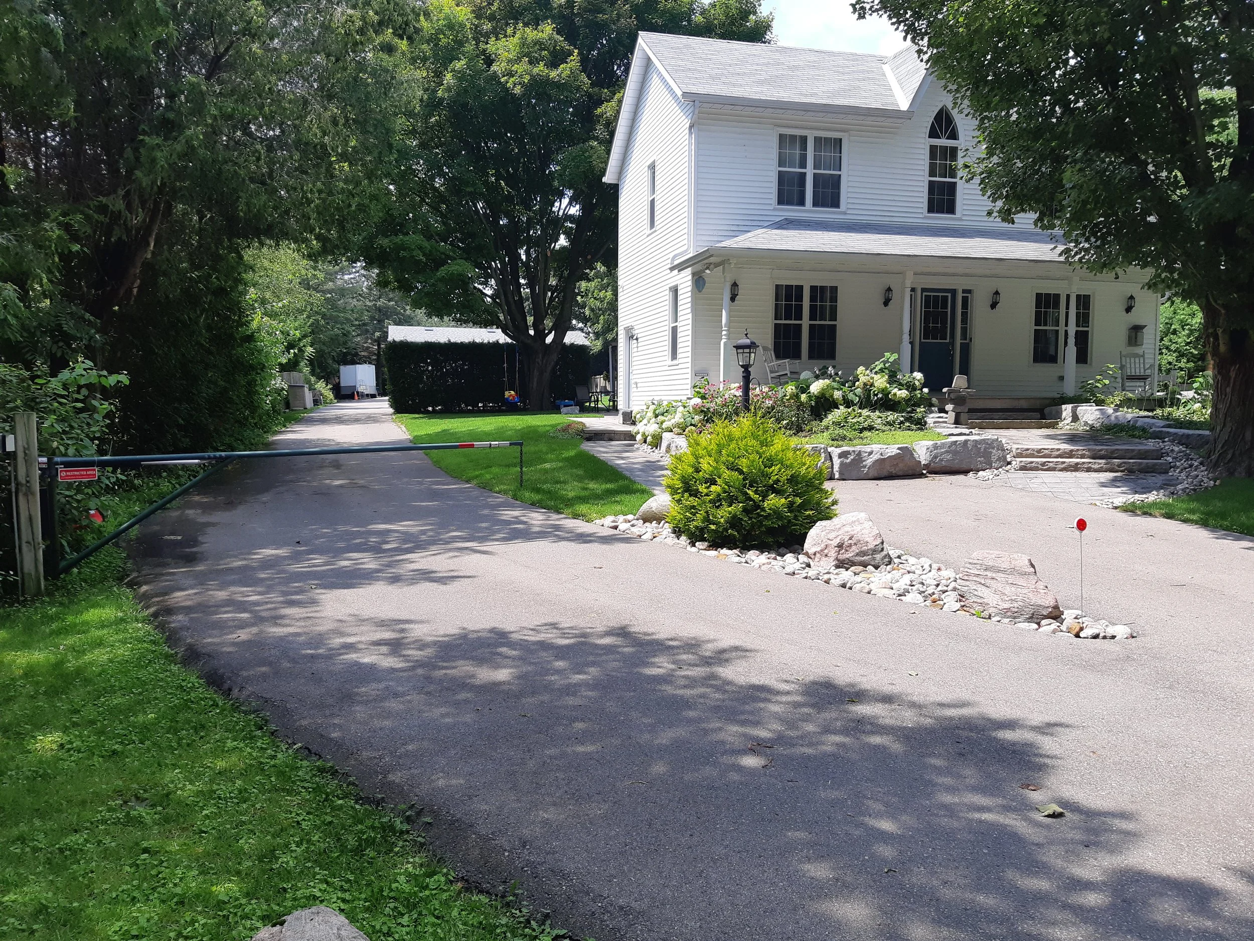 A white two-story house with a porch, surrounded by trees and a garden with white flowers. A driveway leads to the house, with a gate at the entrance.