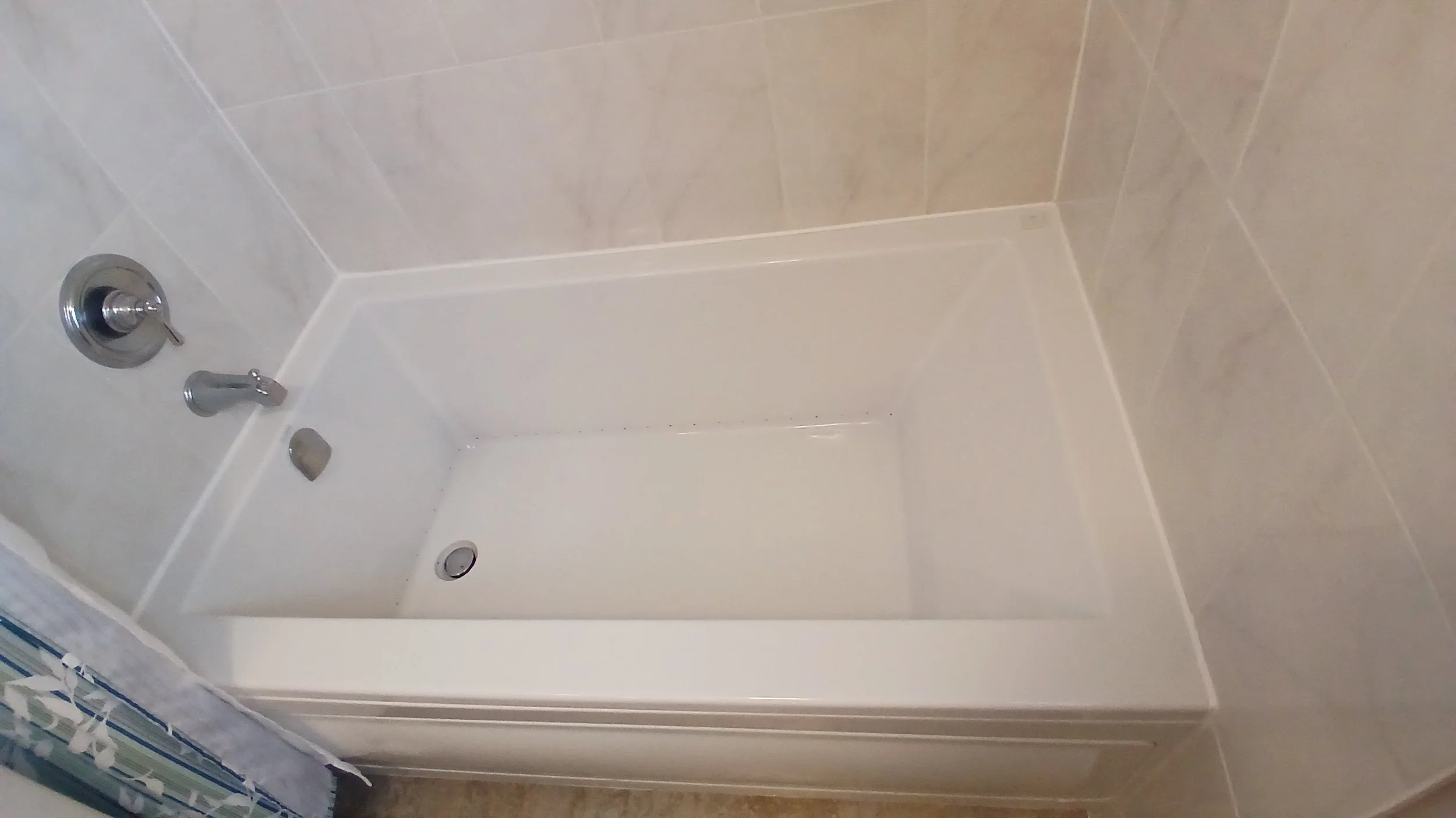 The image shows a white bathtub in a bathroom with beige tiled walls. There are two chrome fixtures, likely a faucet and a knob, on the side of the tub. A shower drain is visible at the bottom of the tub, and part of a shower curtain with a striped p