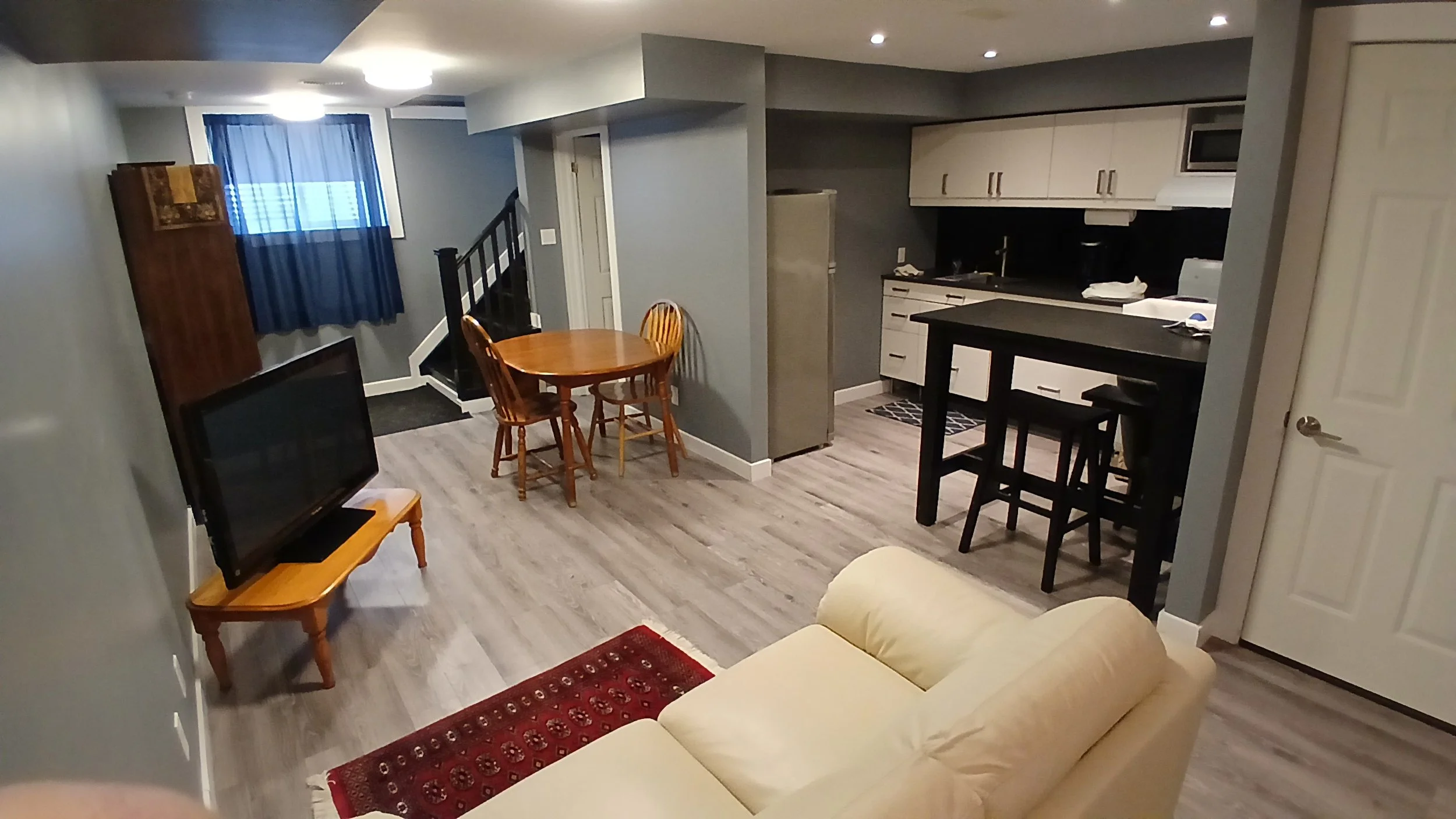 Open-concept living room with a beige sofa, a small wooden table with a TV, a round dining table with four chairs, a kitchen area with white cabinets, black countertops, and a microwave. There is a window with blue curtains and a staircase near the w