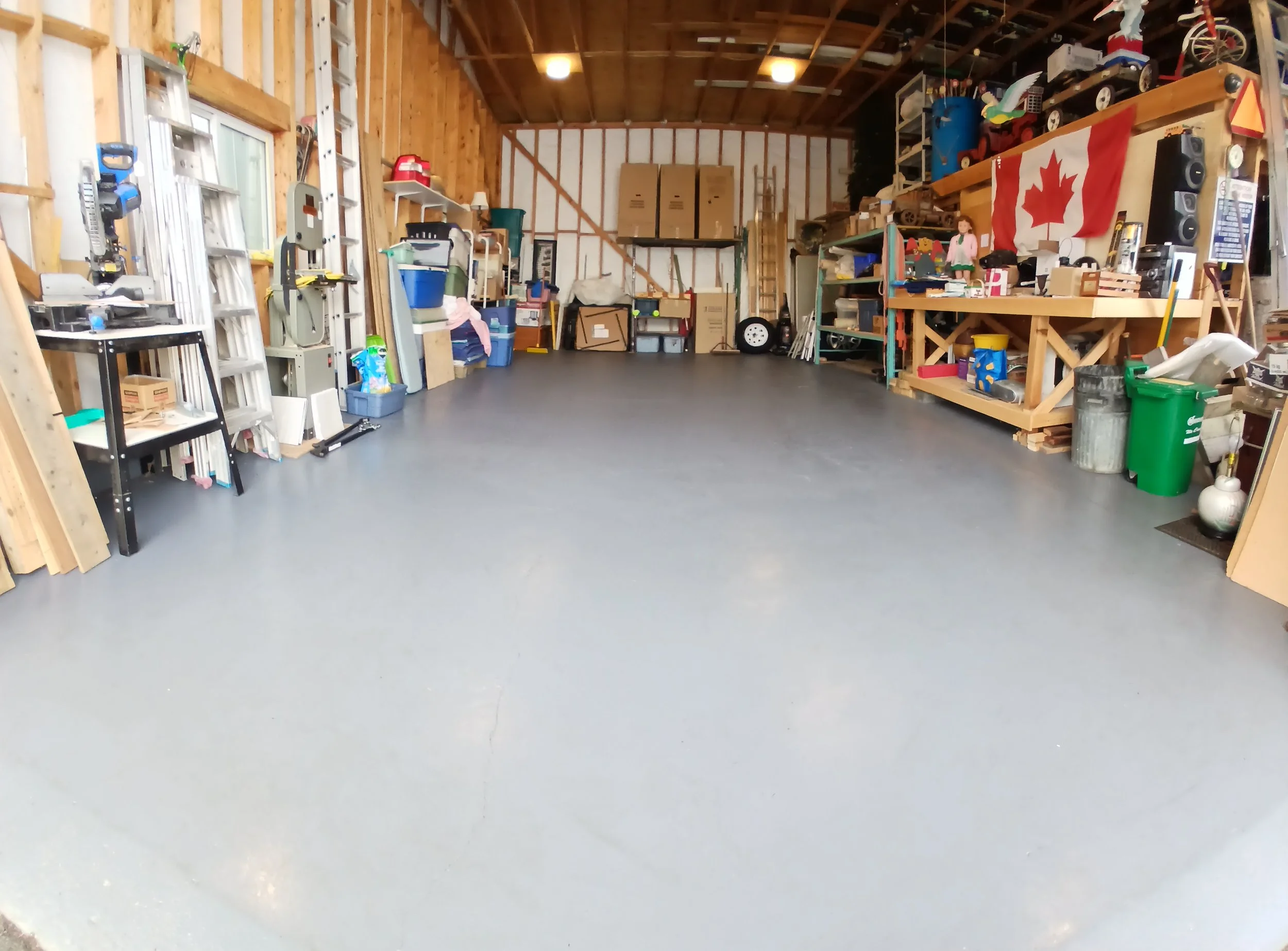 Interior of a garage with wooden walls and shelves filled with various items including toys, tools, and storage bins. A Canadian flag hangs on the right side.