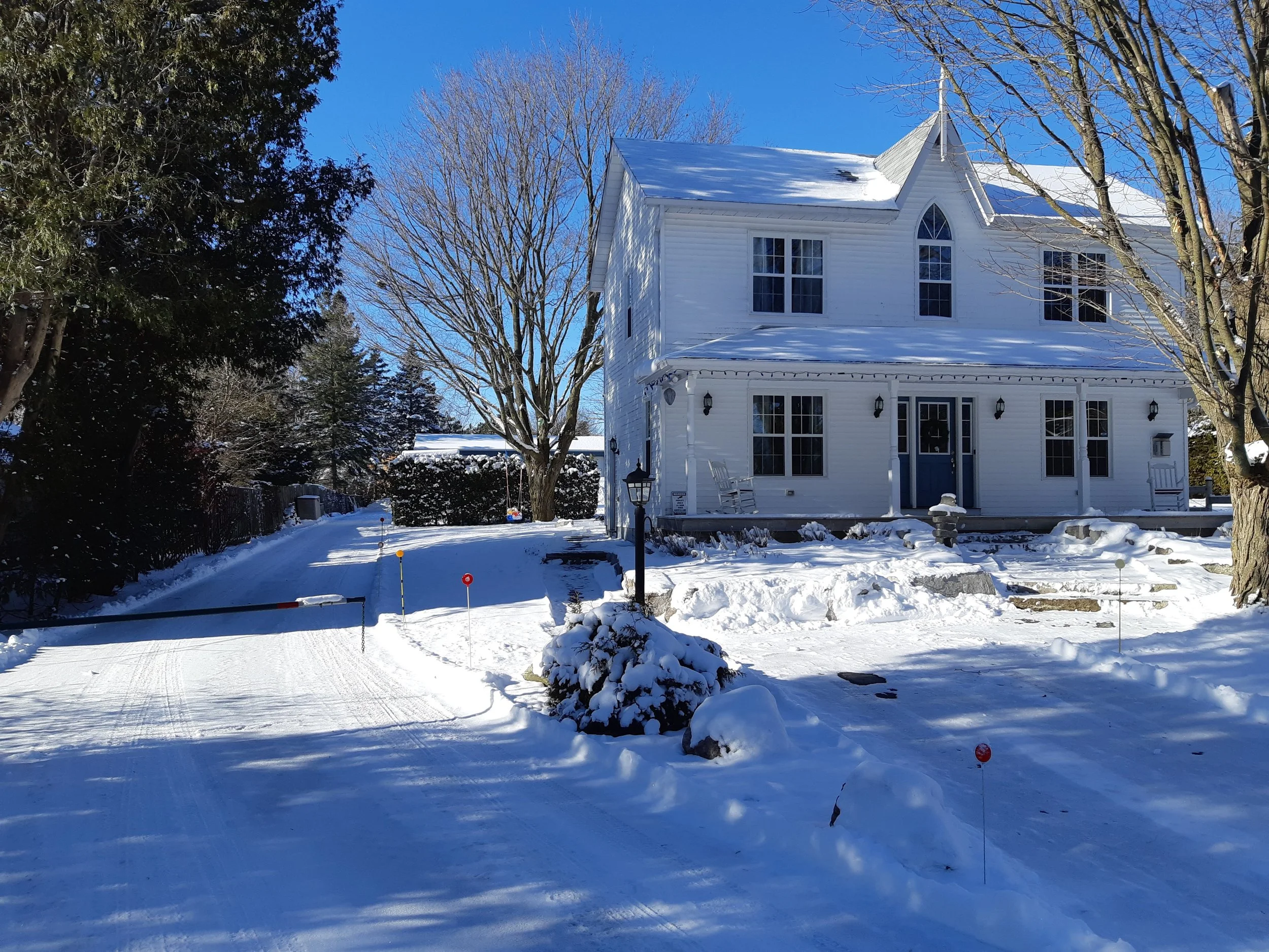White house with snow-covered yard and trees, clear blue sky, and tire tracks in the snow.