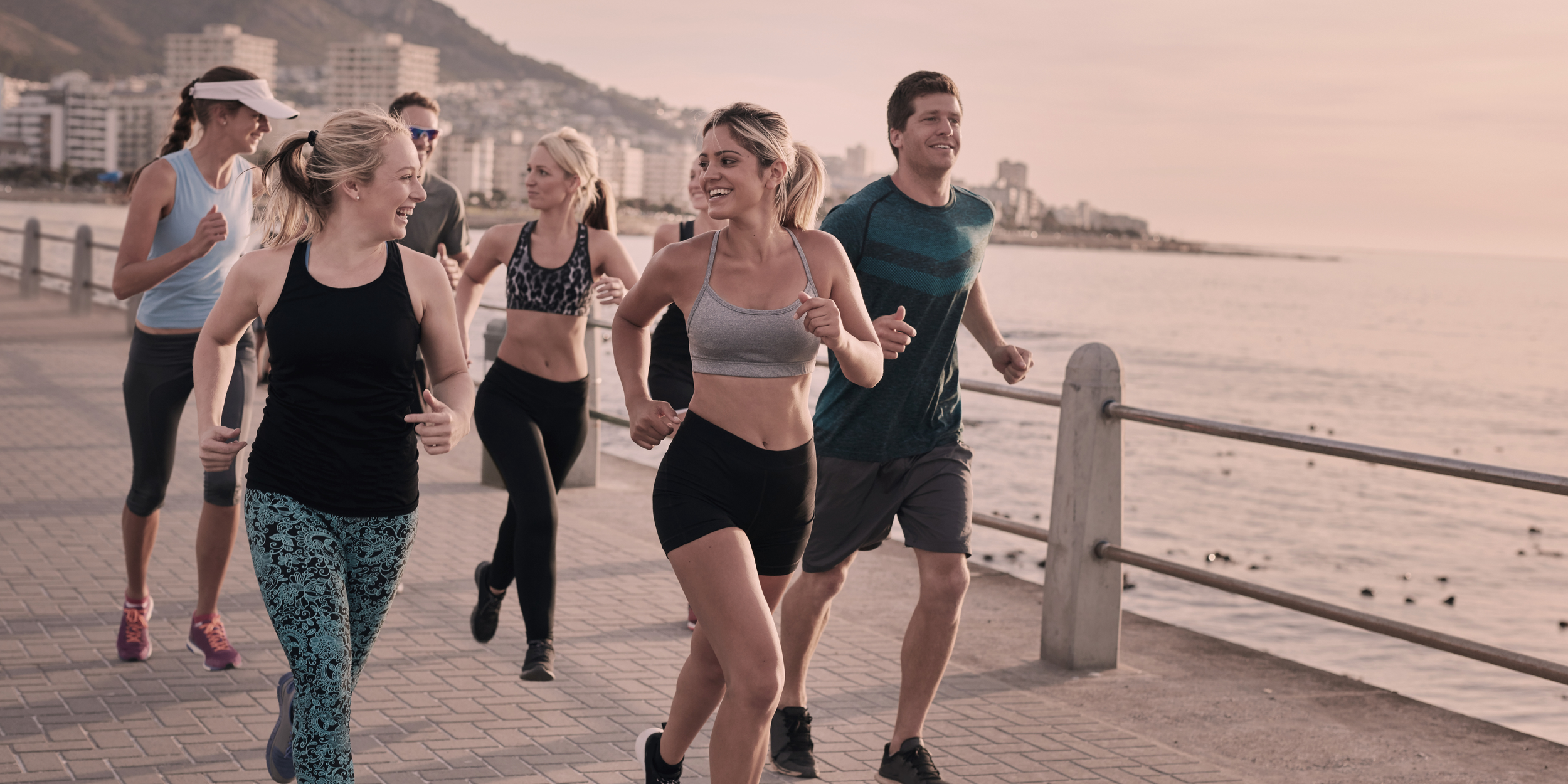 Group of people jogging along a waterfront promenade at sunset, with city buildings and hills in the background.