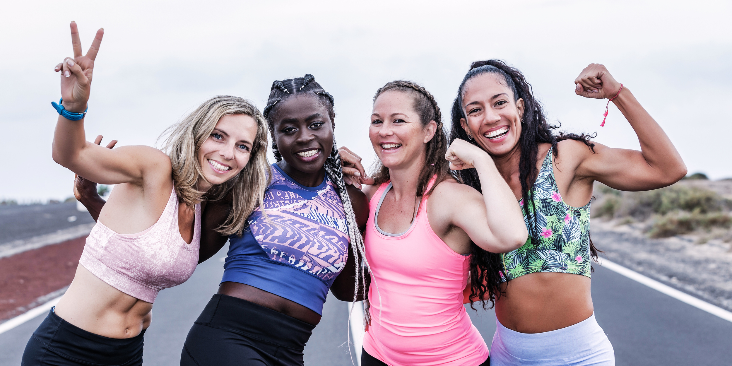 Five diverse women in athletic clothing celebrating together outdoors on a cloudy day, with one showing a victory sign and another flexing her arm.