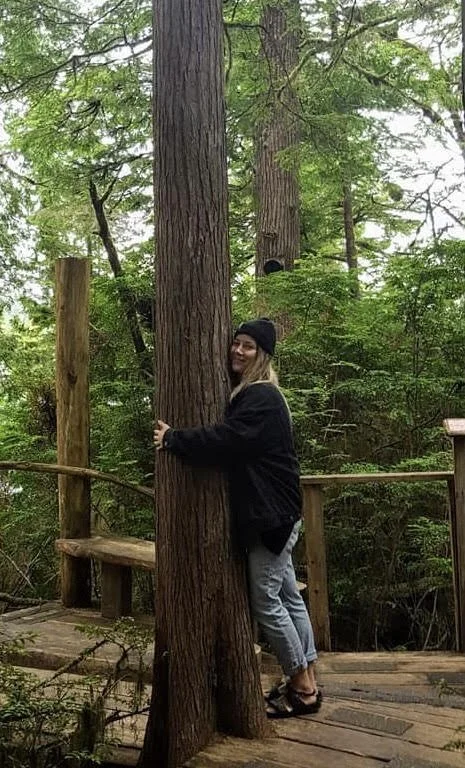 A woman hugging a large tree in a forest, with a forest path and trees in the background.