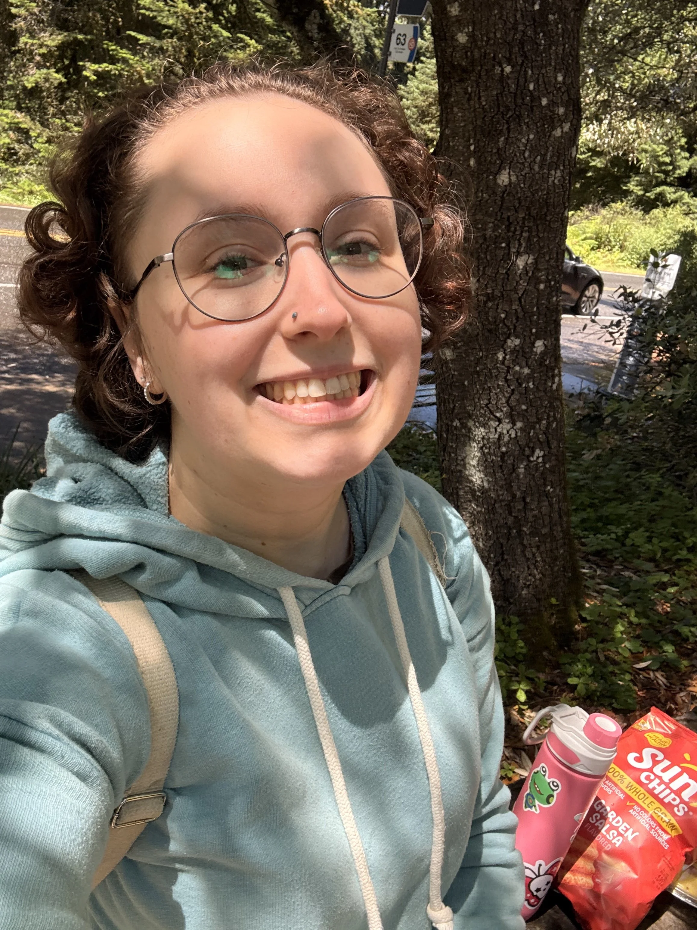 A person with glasses and a nose piercing smiling outdoors in a wooded area, wearing a light teal hoodie with a backpack, standing next to a picnic table with snacks and drinks, including a bag of Sun Chips and a pink insulated water bottle.