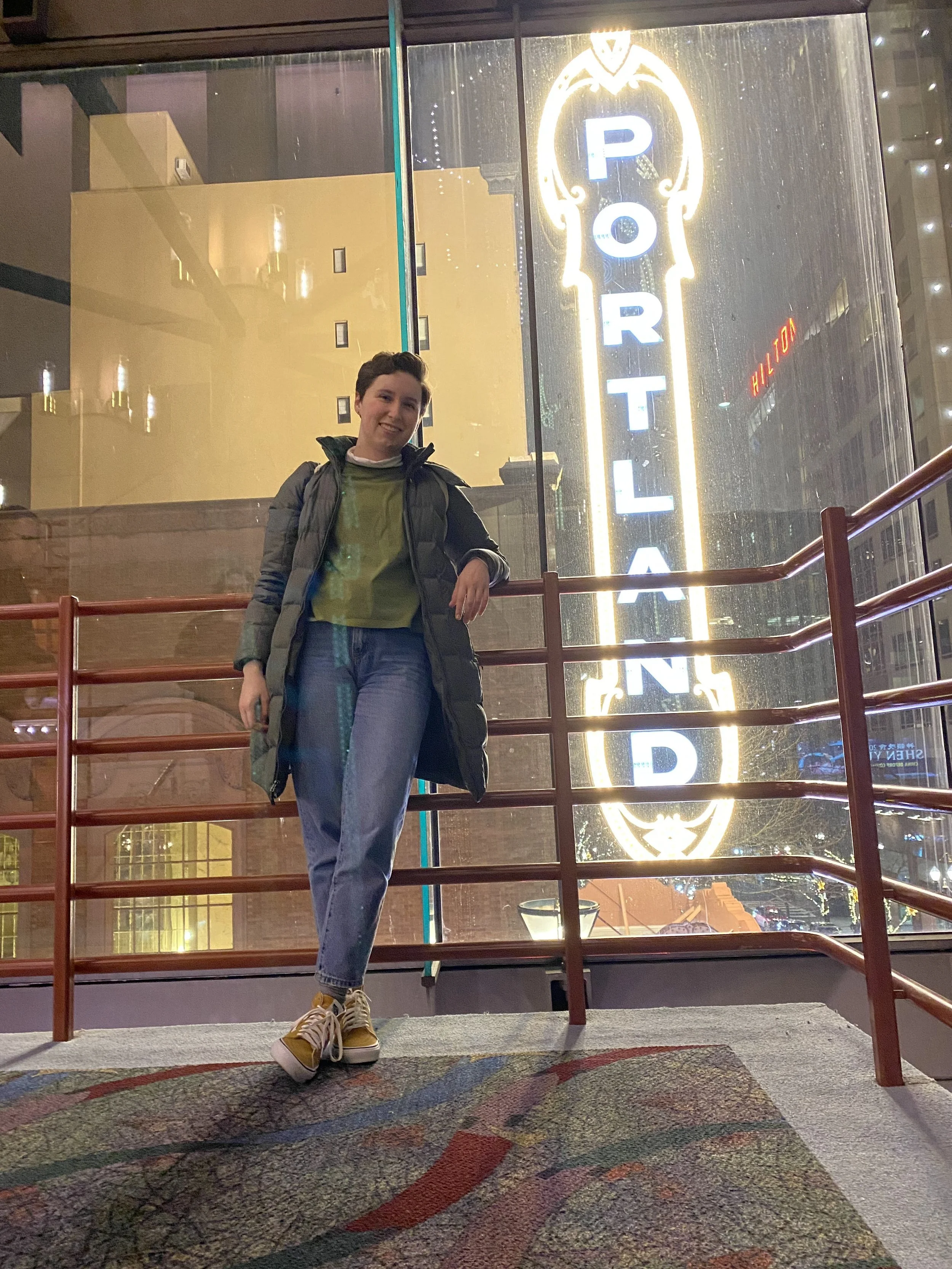 Young person standing in front of a large illuminated sign that reads 'PORTLAND' in a mall or commercial building atrium at night.