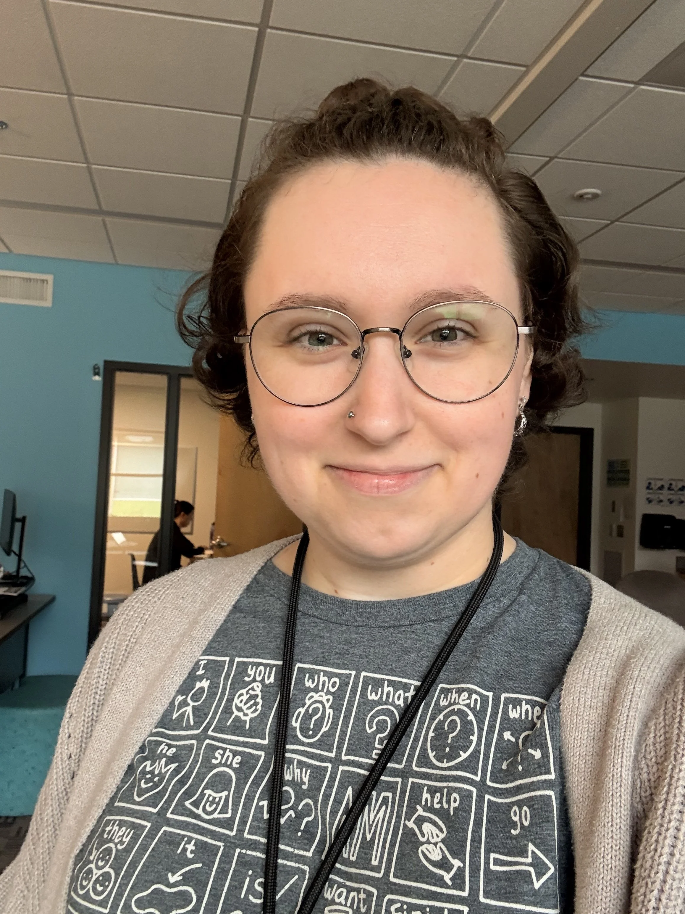 A person with short curly hair wearing glasses, a nose piercing, and a grey graphic T-shirt with AAC device core board visuals on it, standing indoors in a room with a blue wall and door in the background.
