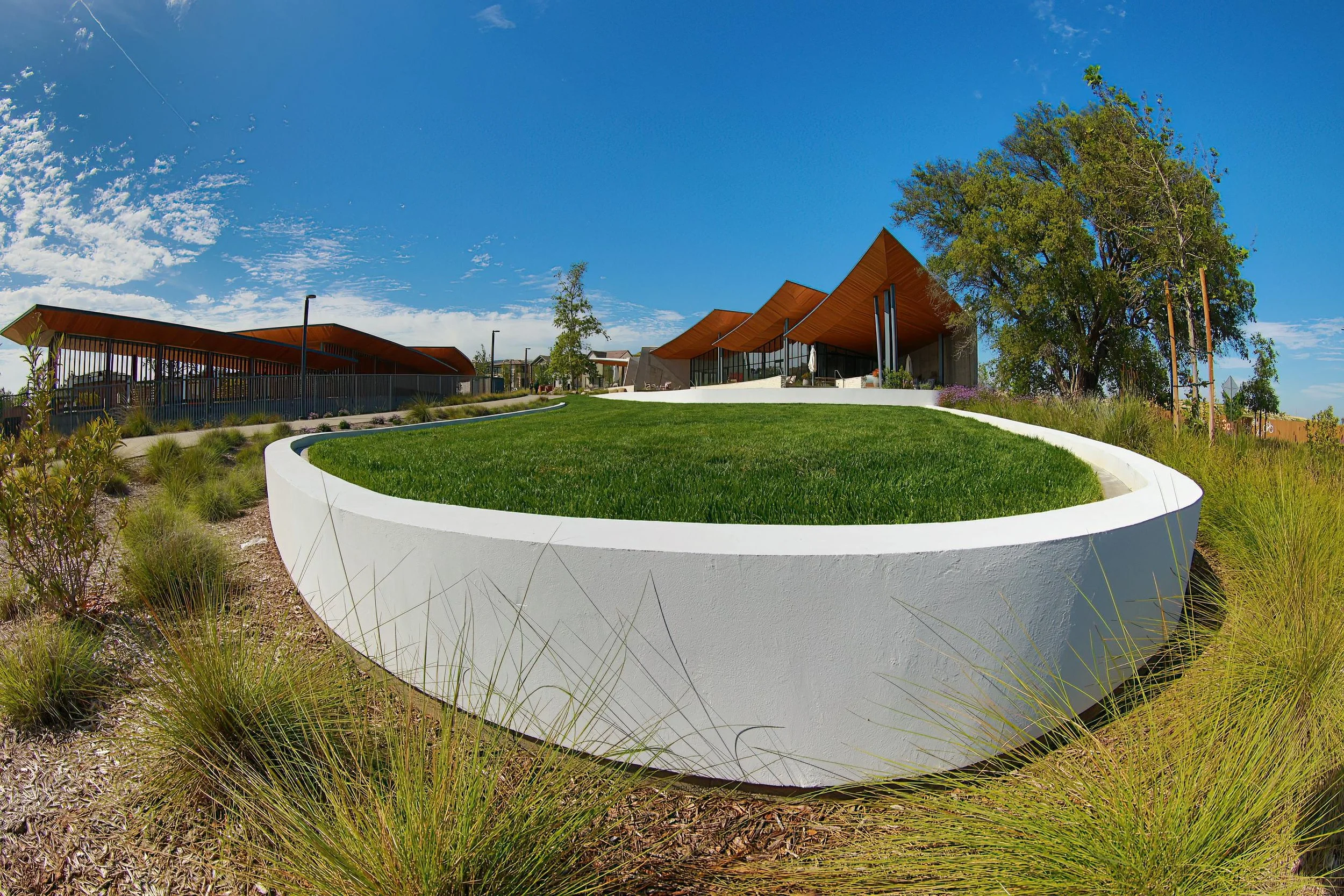Modern architectural building with sloped roofs, surrounded by landscaping including a grassy circular area, plants, and trees, under a partly cloudy blue sky.