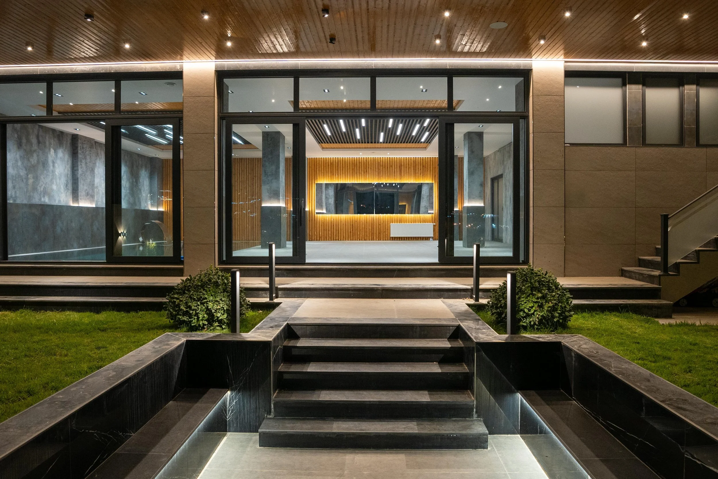 Night view of the entrance to a modern building with glass doors, wooden ceiling panels, warm interior lighting, concrete walls, and a landscaped pathway with stairs and trimmed bushes