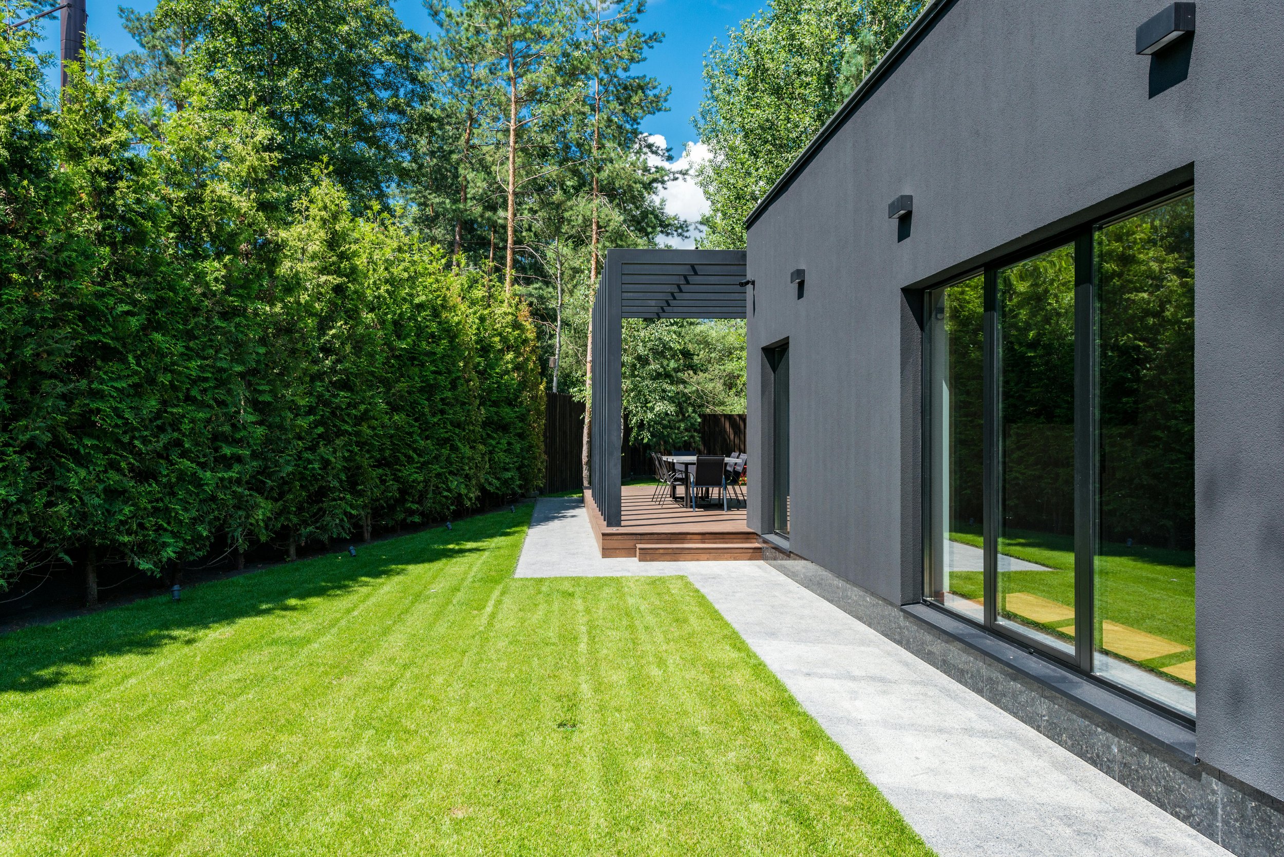 Modern house with large glass sliding doors, a wooden patio with outdoor table and chairs, neatly manicured green lawn, tall green hedge privacy wall, and trees in the background under a bright blue sky.