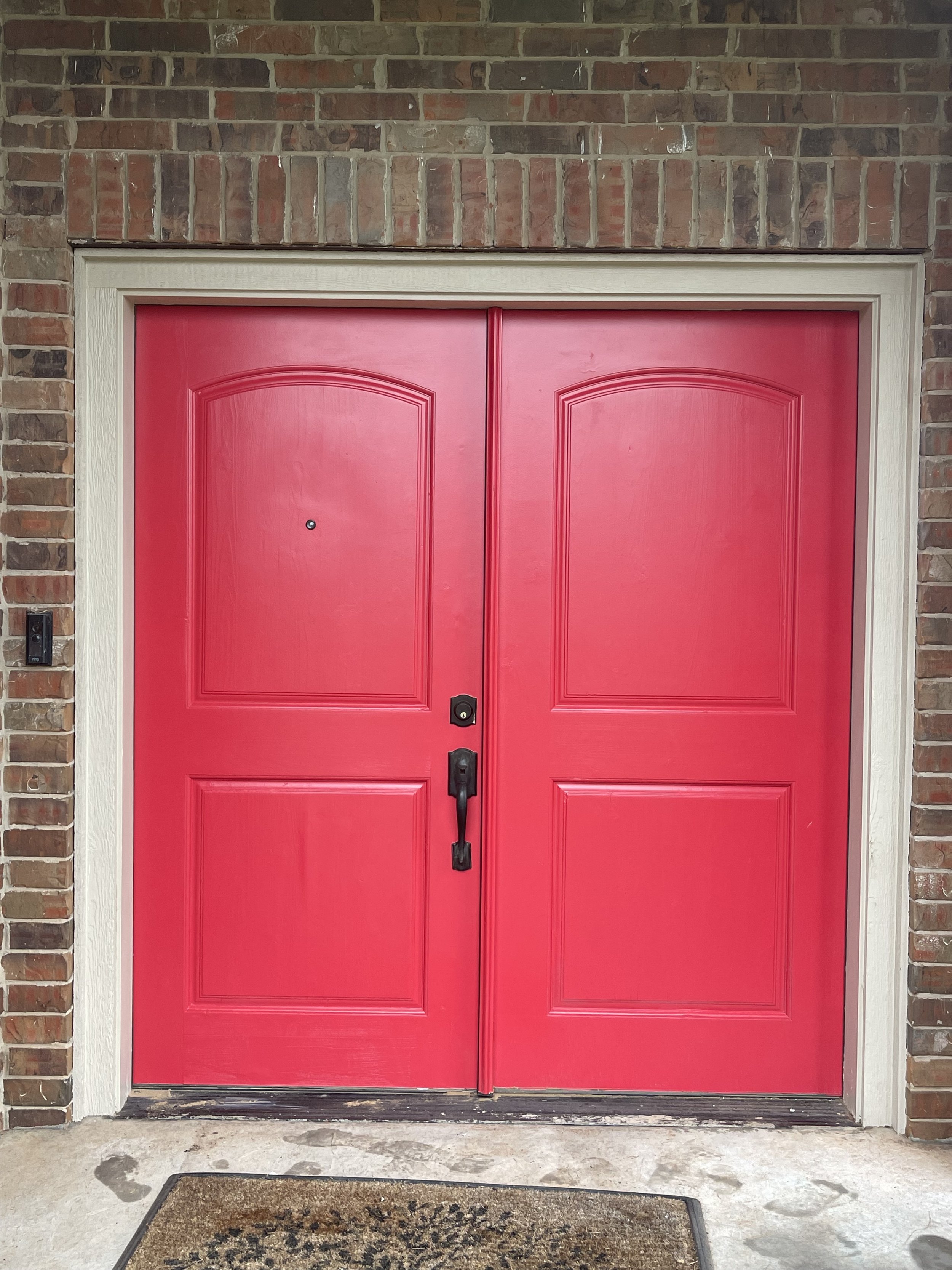 Red double doors with black hardware, set in a brick wall, with a doormat in front on concrete ground.