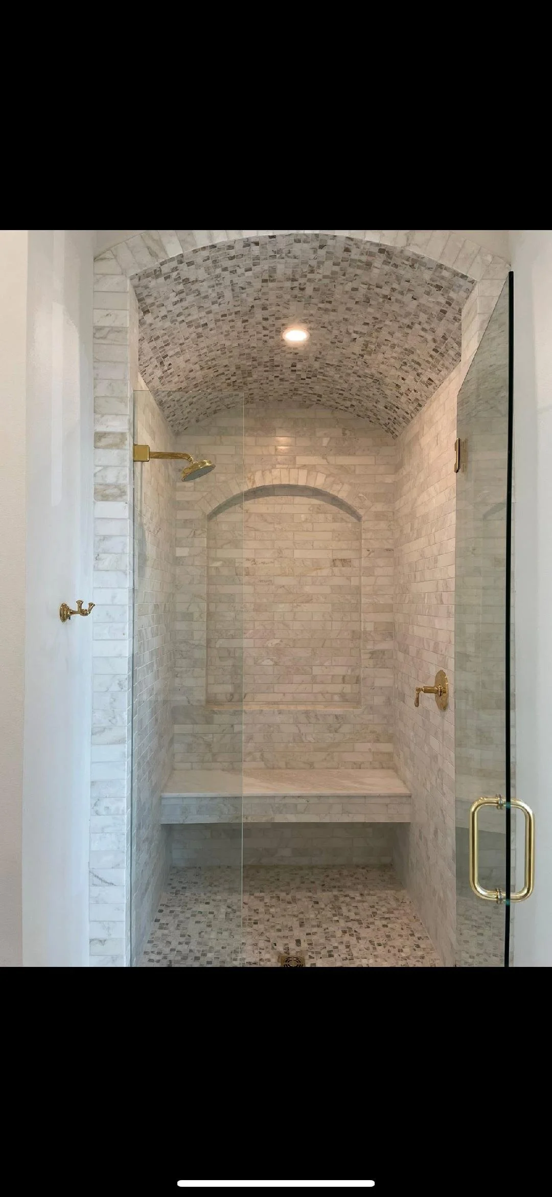 A walk-in shower with a glass door, marble brick walls, a ceiling arch with mosaic tiles, a built-in bench, gold fixtures, and a recessed ceiling light.