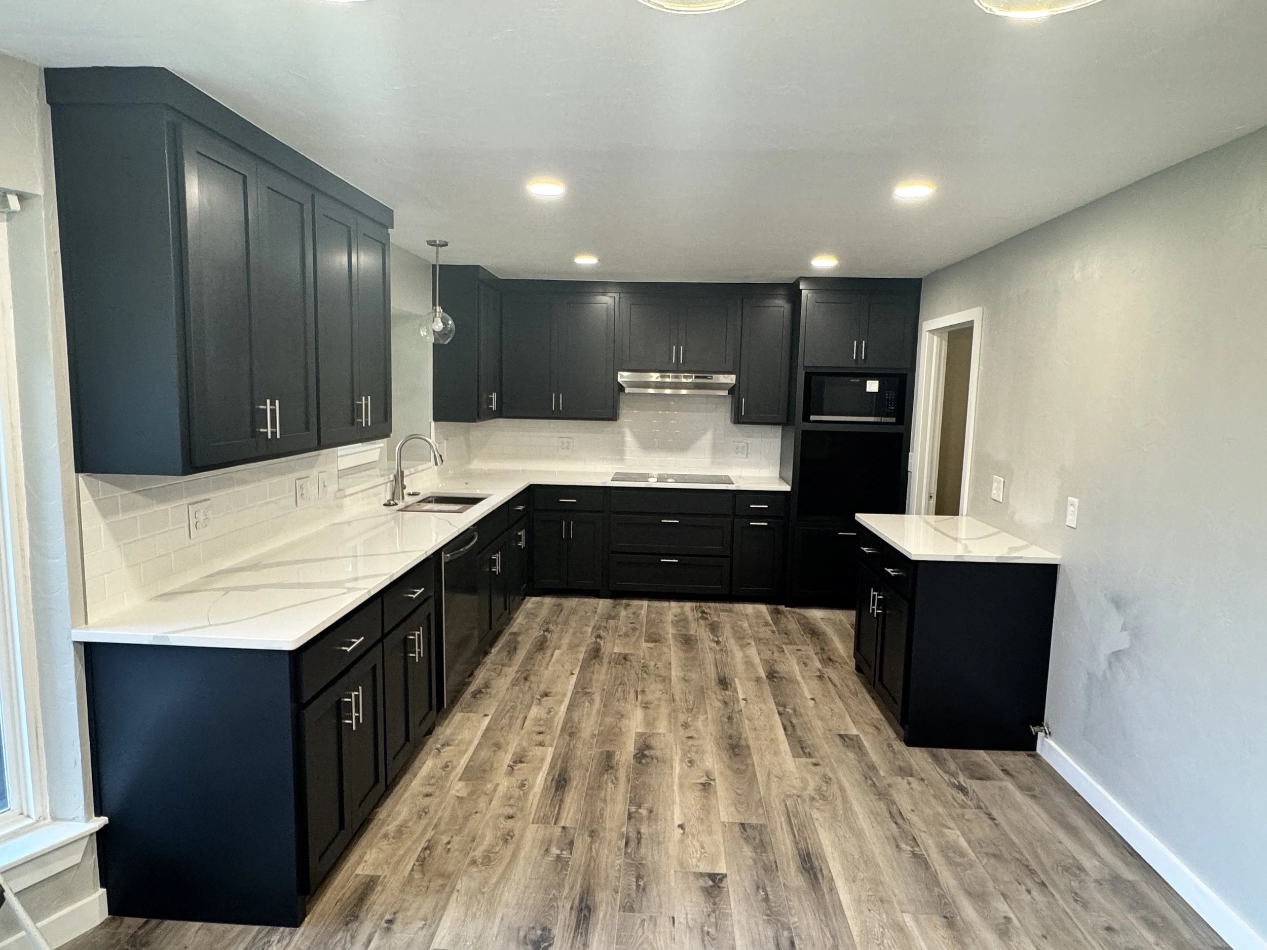 A modern kitchen with black cabinets, white countertops, a white subway tile backsplash, and a dark wood floor. There is a sink with a modern faucet, and a microwave oven built into the cabinetry. Recessed lighting illuminates the room.