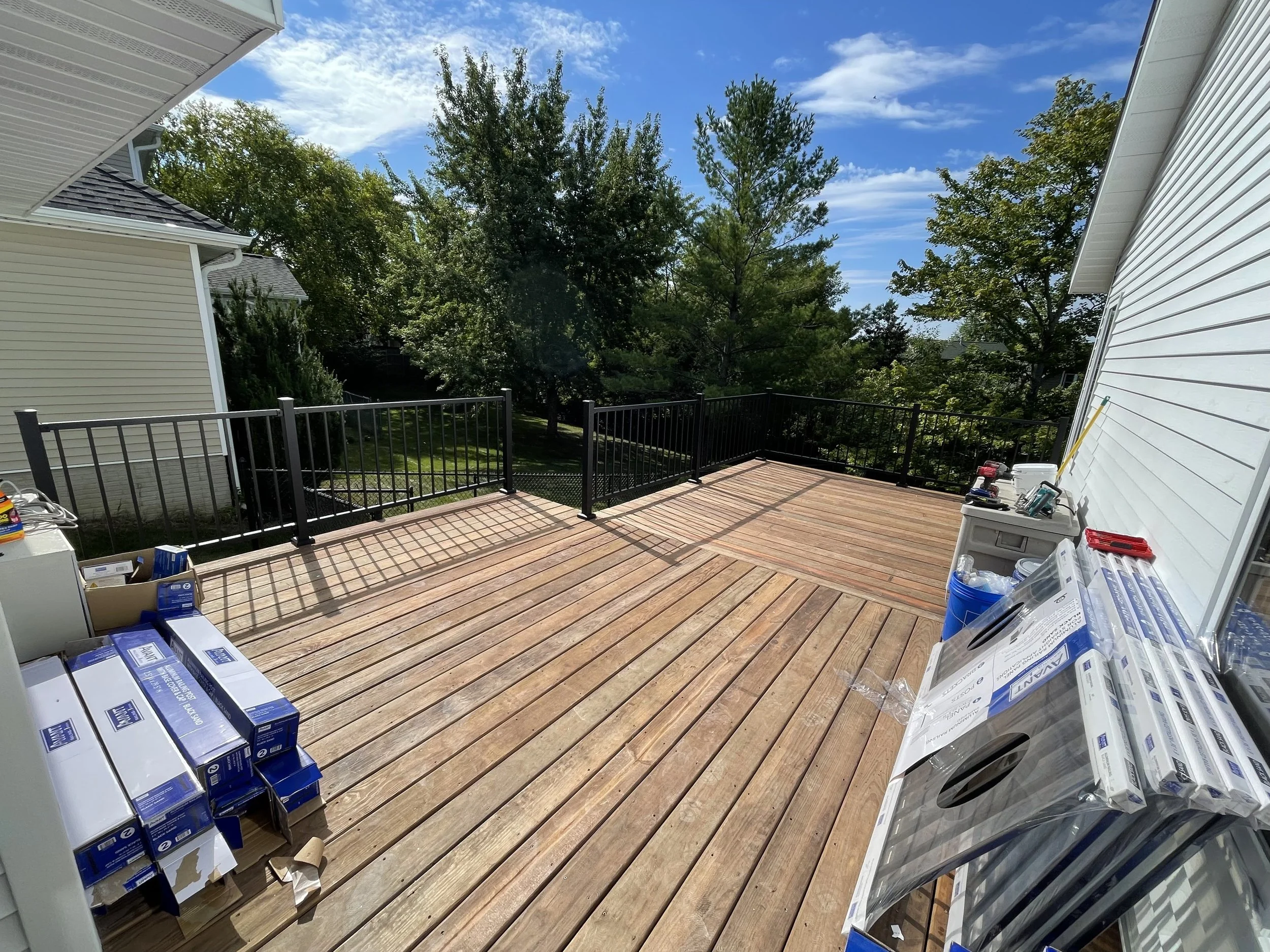A spacious wooden deck with building siding on either side, a black railing around the perimeter, and trees in the background under a partly cloudy blue sky. There are boxes and tools on the deck, indicating ongoing work.