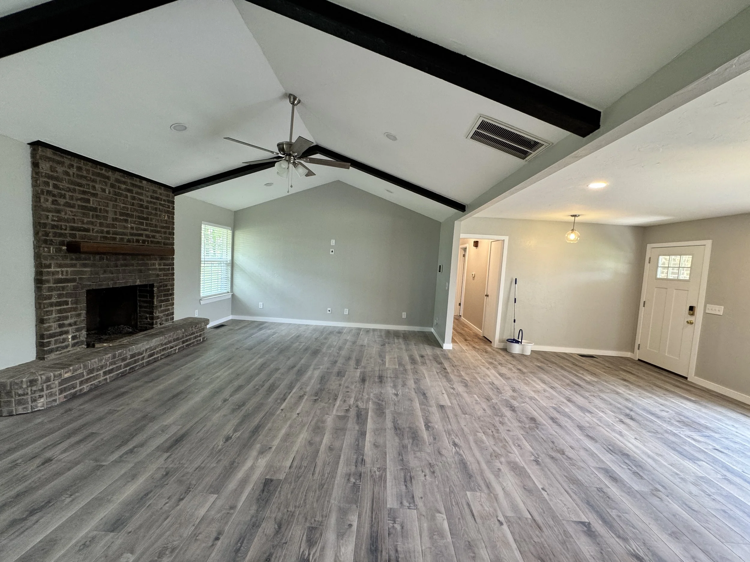 Open-concept living room with wood-look flooring, a brick fireplace, a ceiling fan, and neutral-colored walls. Large windows let in natural light.