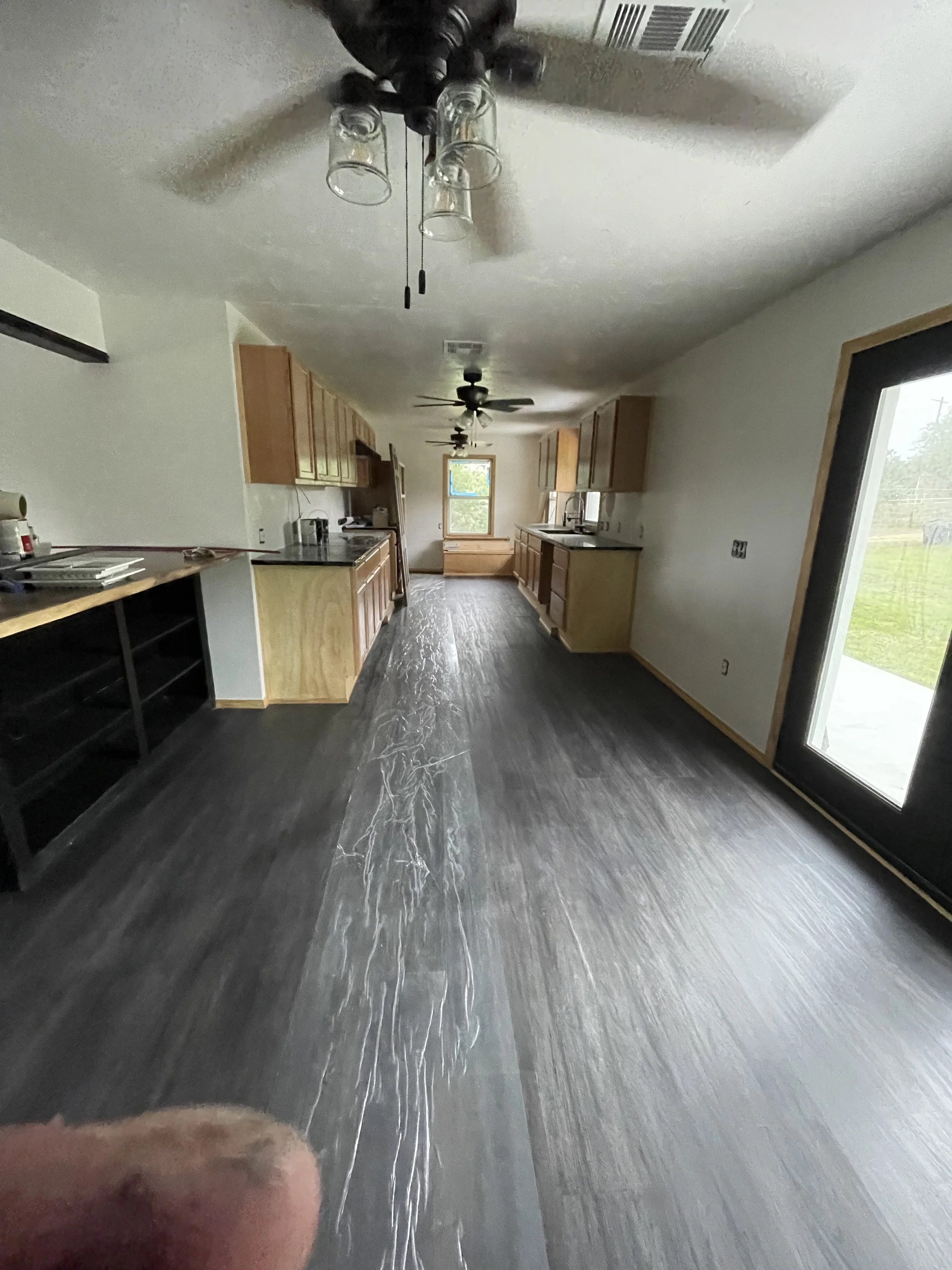 A kitchen with wooden cabinets, a window, black countertops, and a sliding glass door with a screen. The ceiling has two black ceiling fans with lights, and the floor appears to be dark wood or laminate.