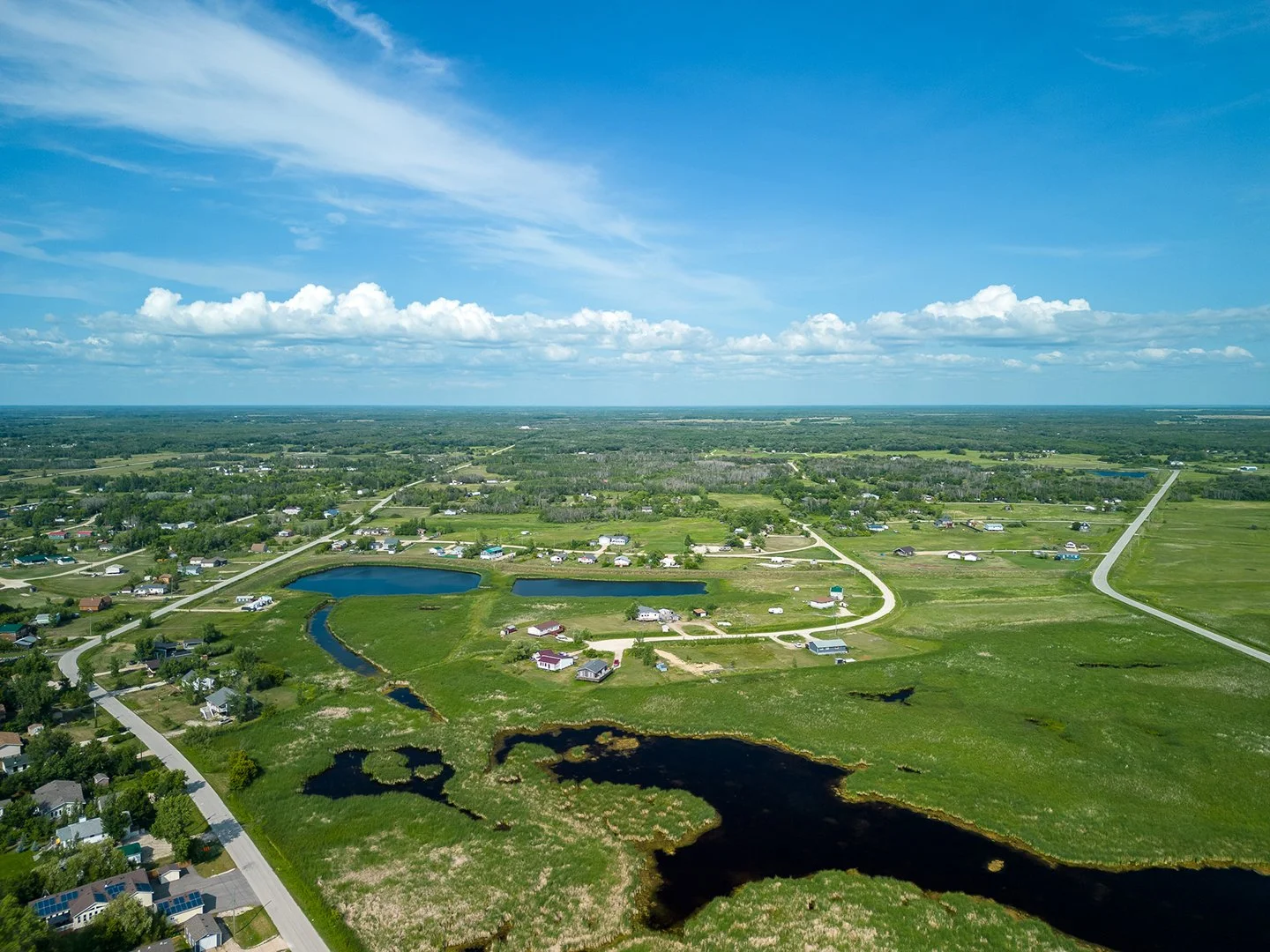 Aerial view of a rural landscape with ponds, green fields, houses, and roads under a blue sky with scattered clouds.