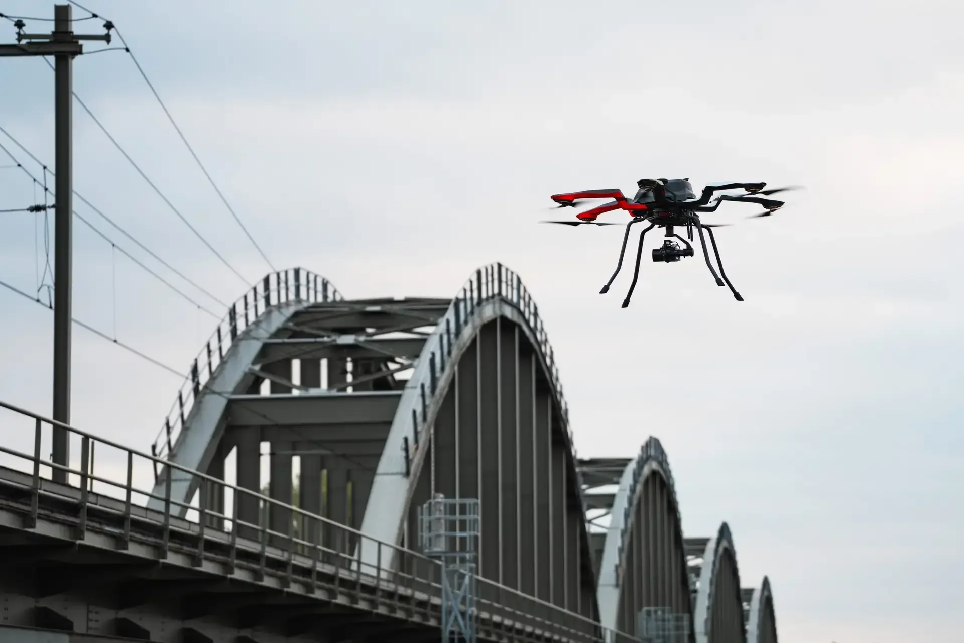 A drone flying over a bridge with arched supports and a railway track in the background.