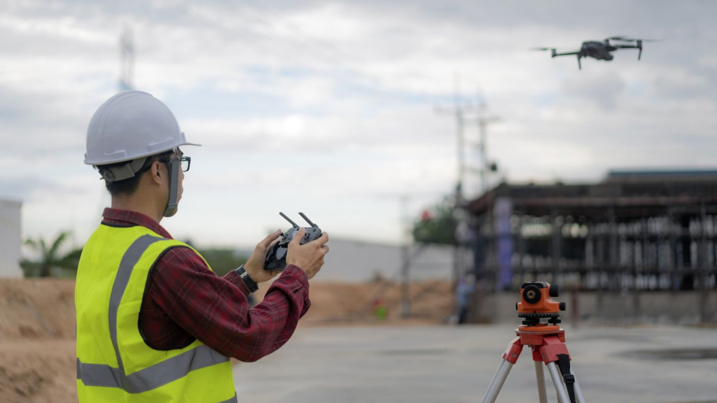 A construction worker wearing a white hard hat and yellow safety vest operates a drone using a remote control at an outdoor construction site. A surveying instrument is set up on a tripod nearby, with a cloudy sky in the background.