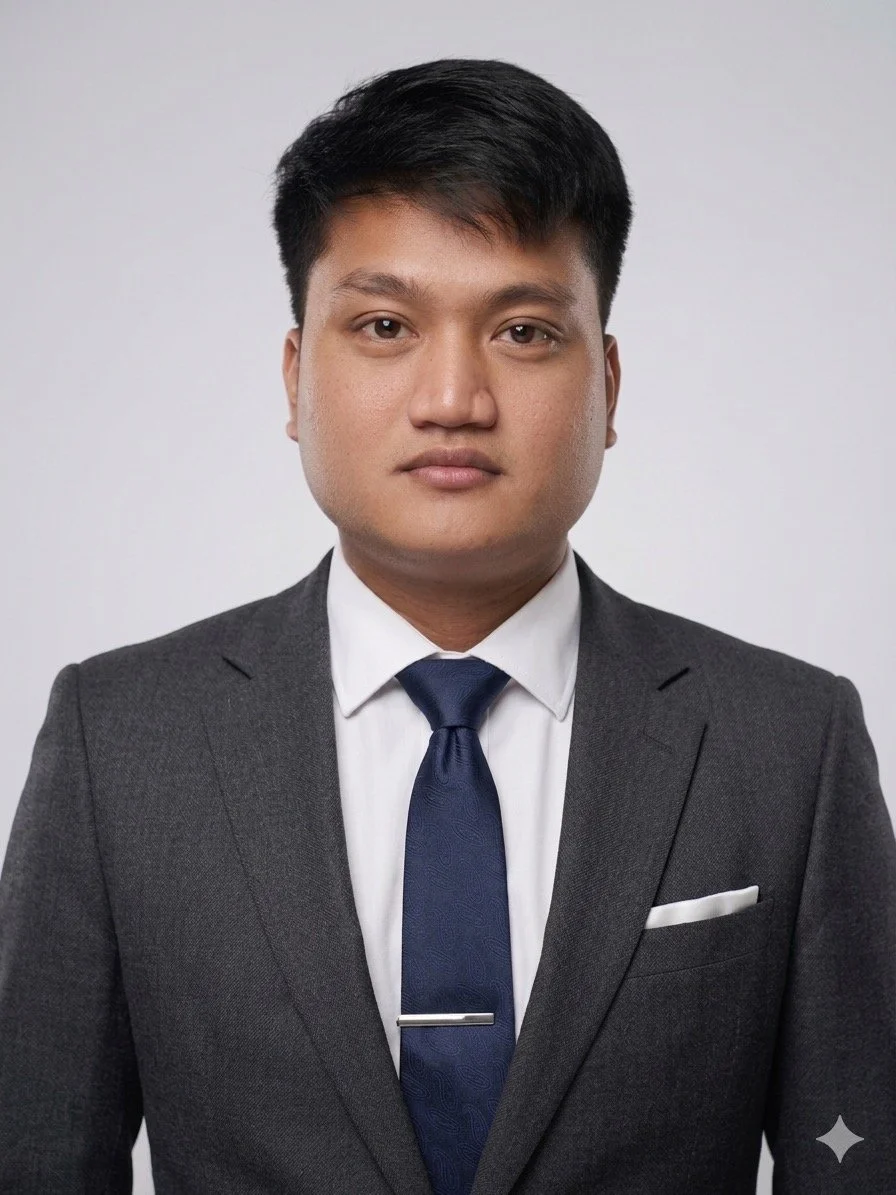 A young man in a dark gray suit, white dress shirt, and navy tie, standing against a plain light background.