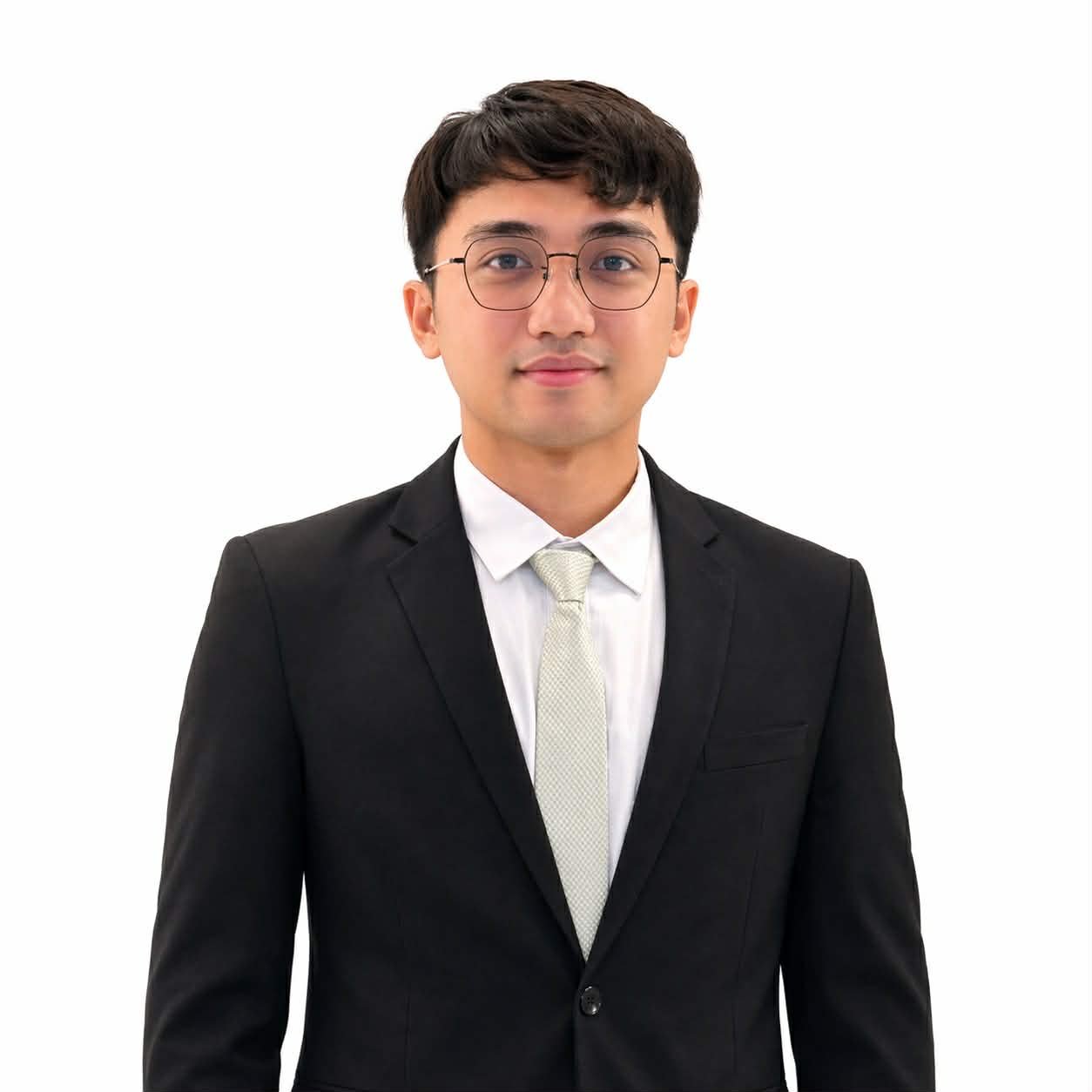 Young man in a black suit, white shirt, cream tie, glasses, smiling against a white background.