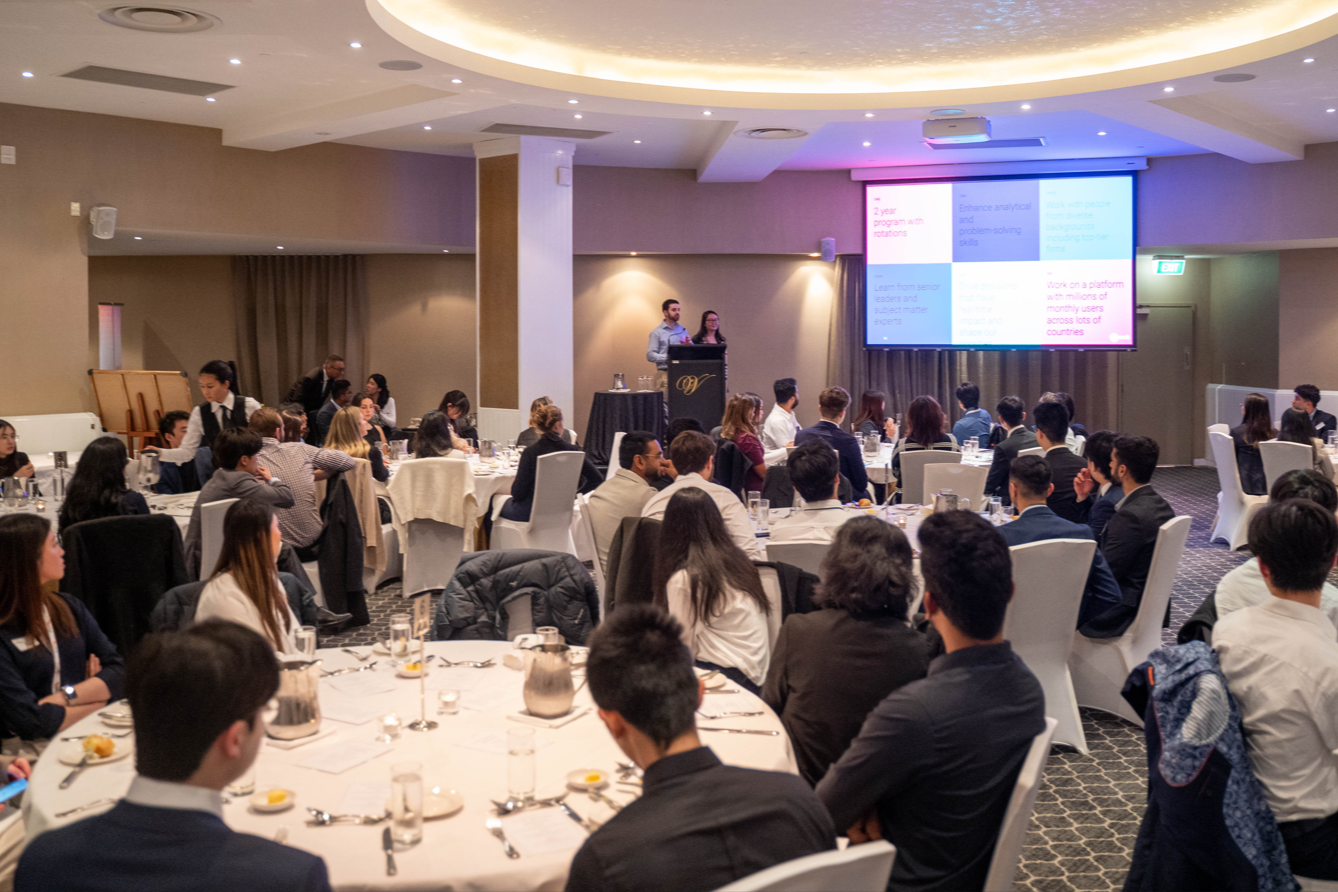 A conference room filled with round tables of attendees listening to a presentation on a large screen. Two speakers stand at a podium in front of the screen.