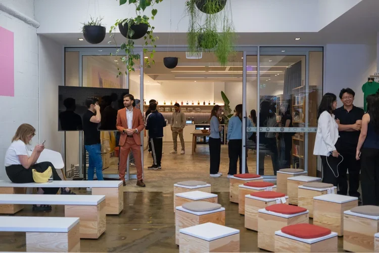 People socializing inside a modern, open space with hanging plants, benches with cushions, and a glass entrance, possibly a lounge or coworking area.